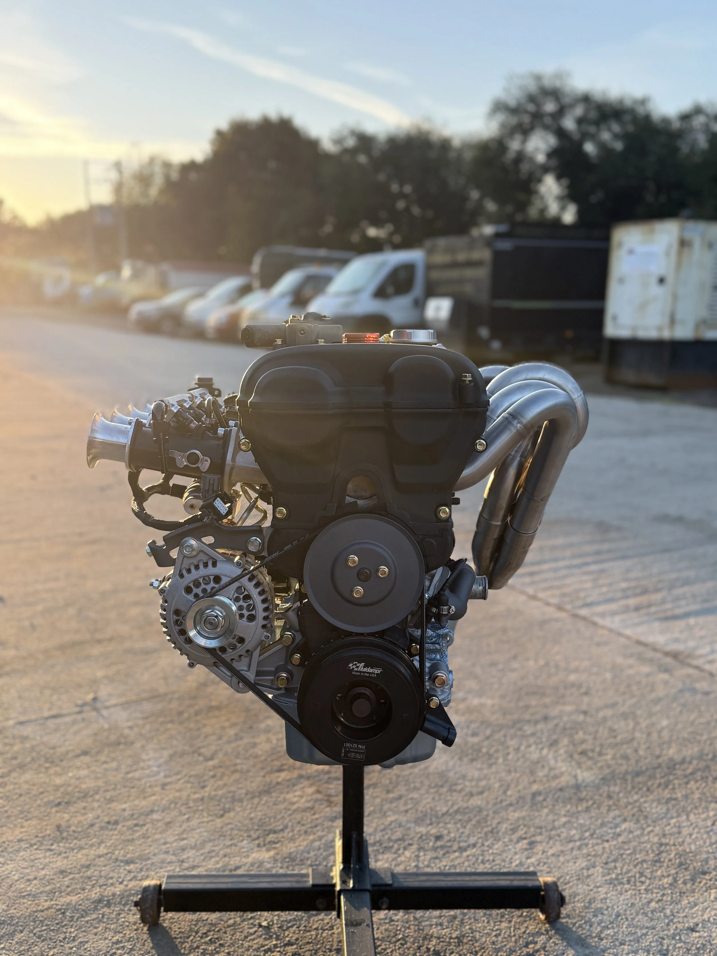 A car engine mounted on a stand in an outdoor parking lot during sunset. NA ITB Mazda MX5 Miata Engine 1.8