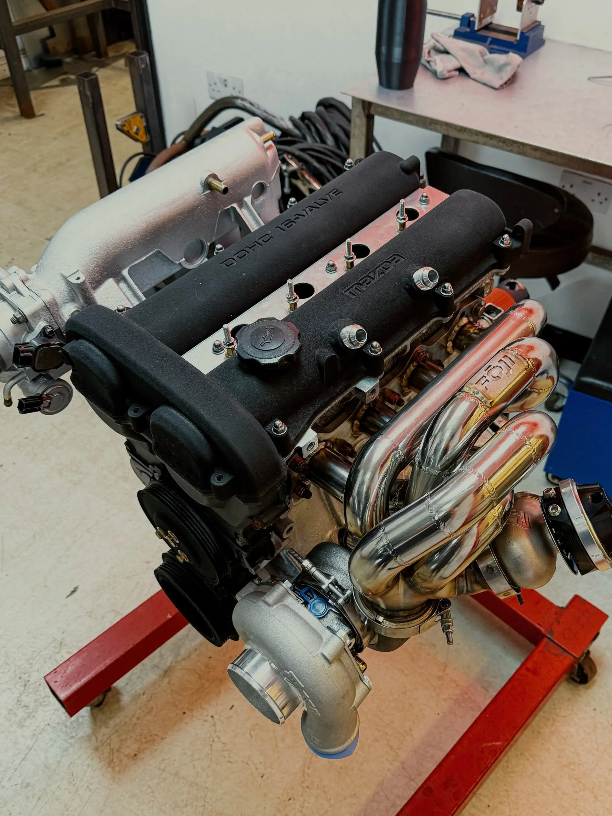 A car engine mounted on a red stand in a workshop. 1.6 Mazda Mx5 engine with a turbo manifold mounted