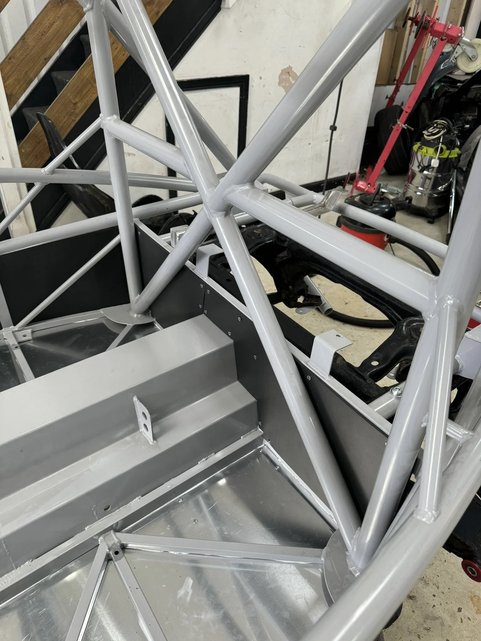 Partially assembled race car chassis with a roll cage and metal panels in a workshop.