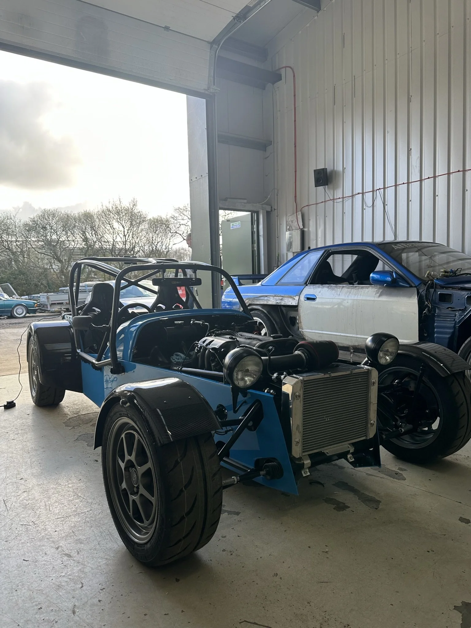 An open-wheel race car with a visible engine and radiator, parked inside a garage with a partially assembled car in the background. MK Sports Kit car