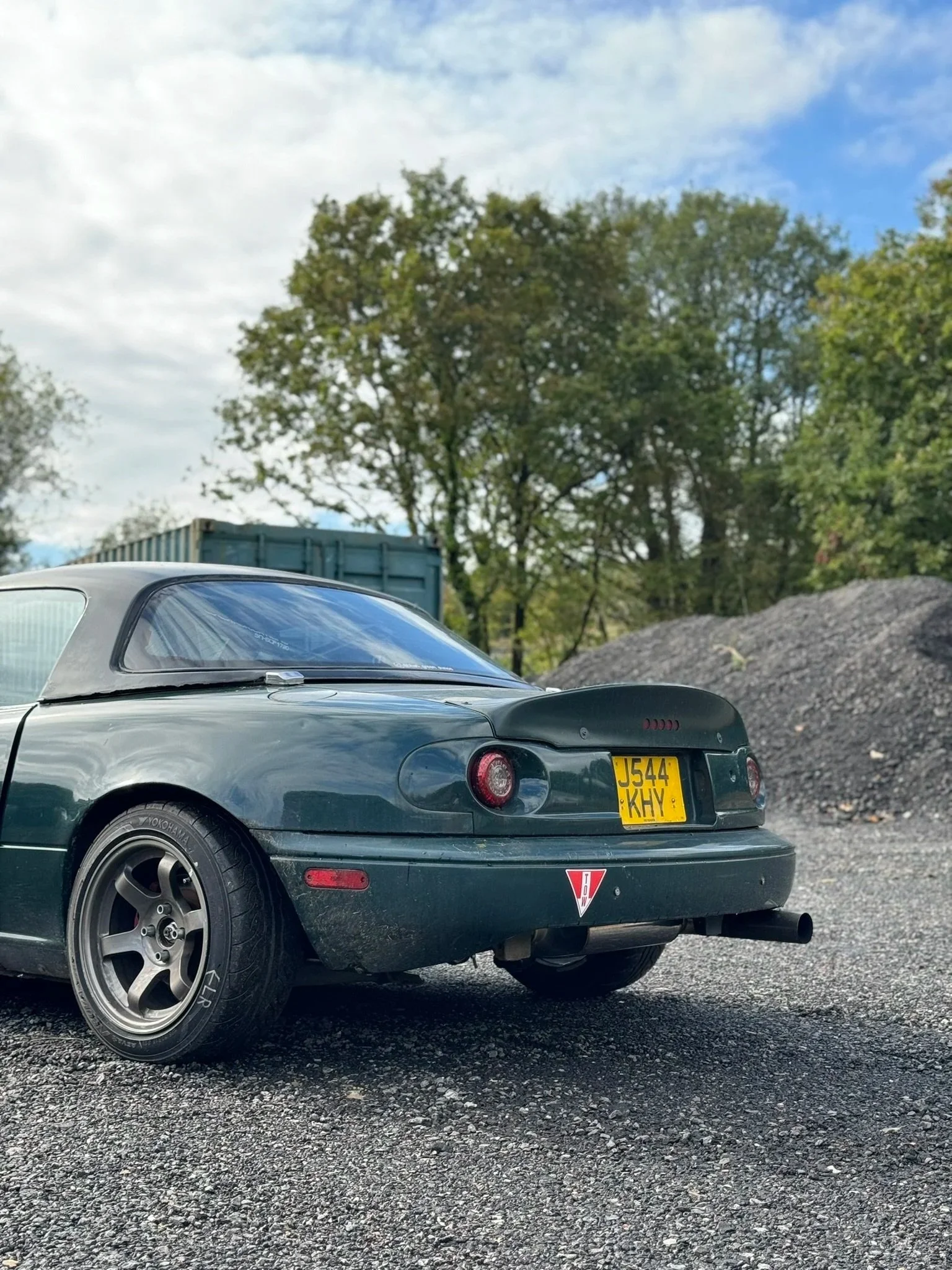 Rear view of a dark green Mazda MX-5 Miata car parked on gravel with trees and a blue sky in the background.
