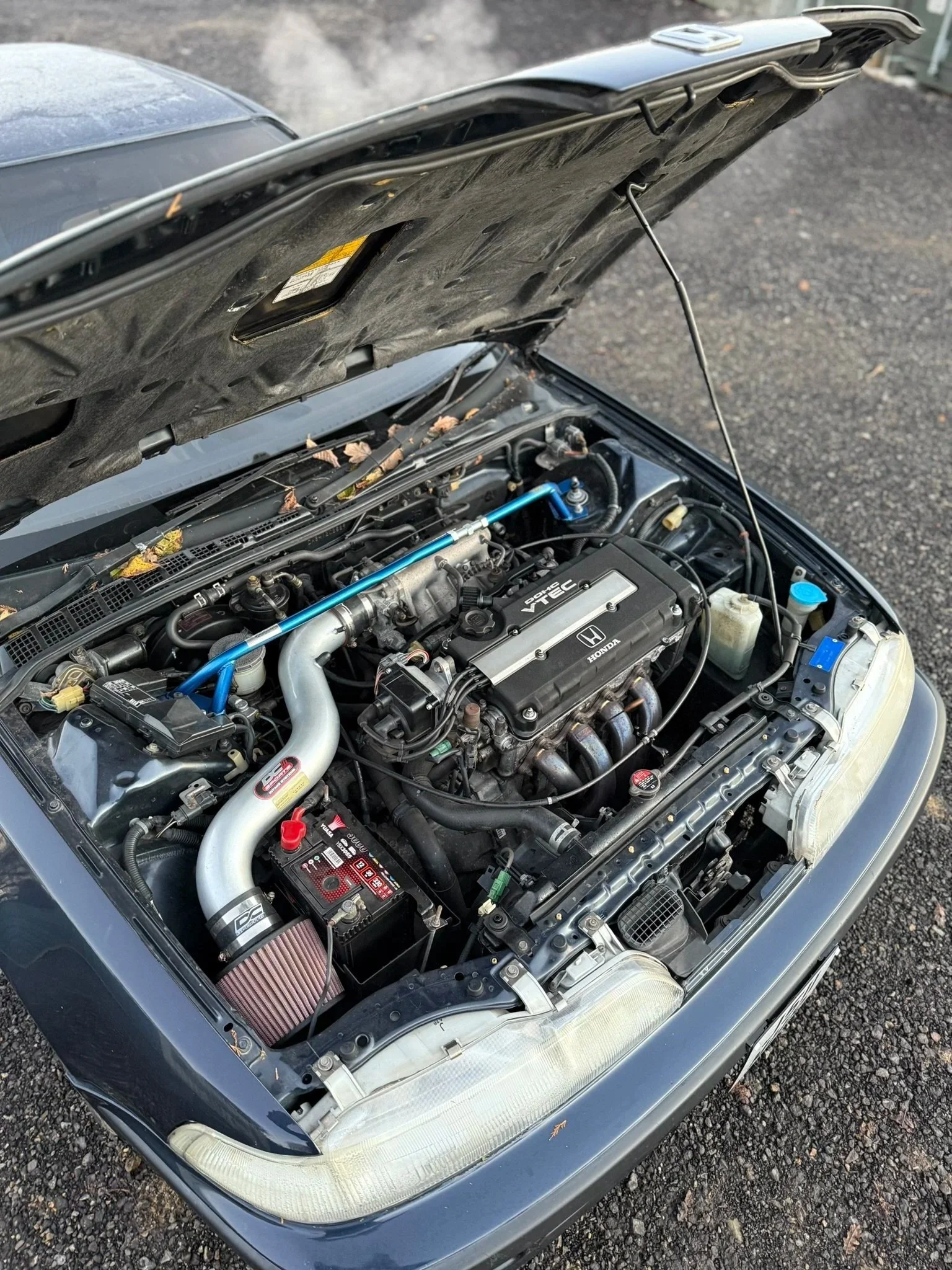 Open hood of a car showing a Honda engine with various components, including a roll cage, battery, air filter, and radiator, parked on a gravel surface. Honda integra DA6