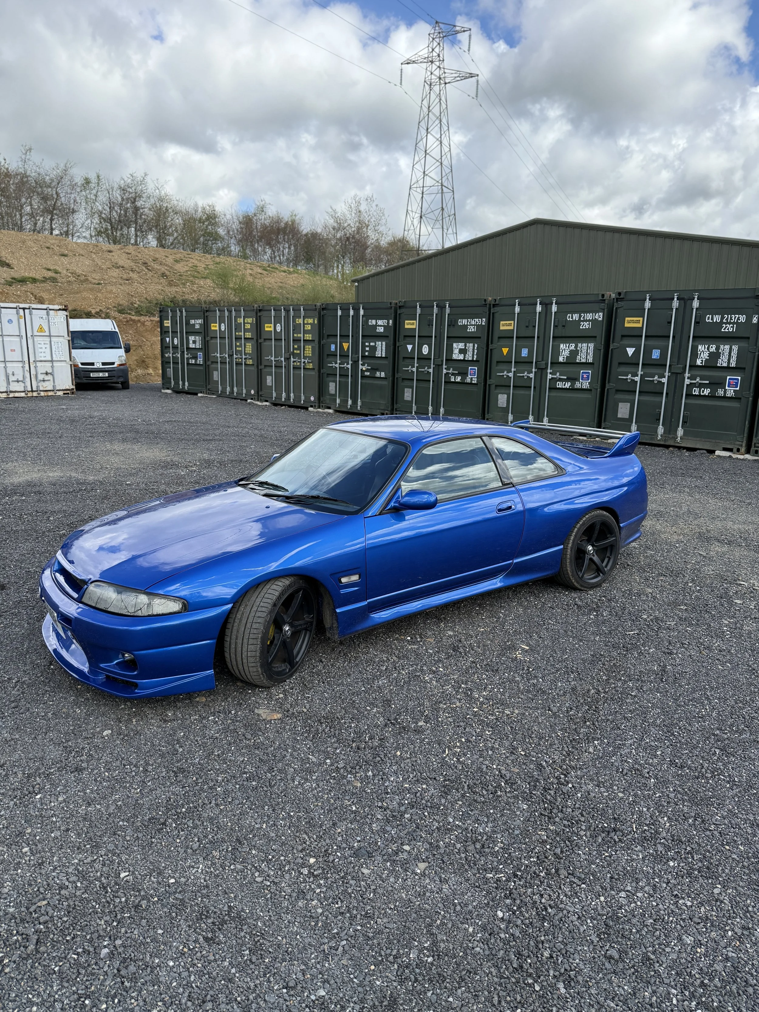 Nissan Skyline r33
A blue sports car with a rear spoiler parked on gravel in front of green shipping containers and a green building under a partly cloudy sky.
