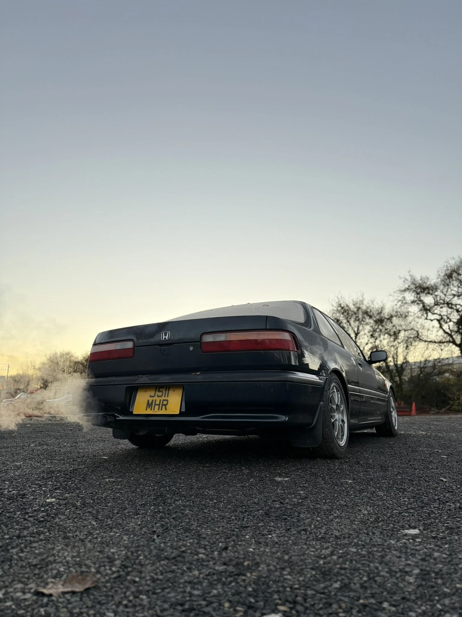 Rear view of a black Honda Integra DA parked on a paved surface at dusk, with trees and orange cones in the background.