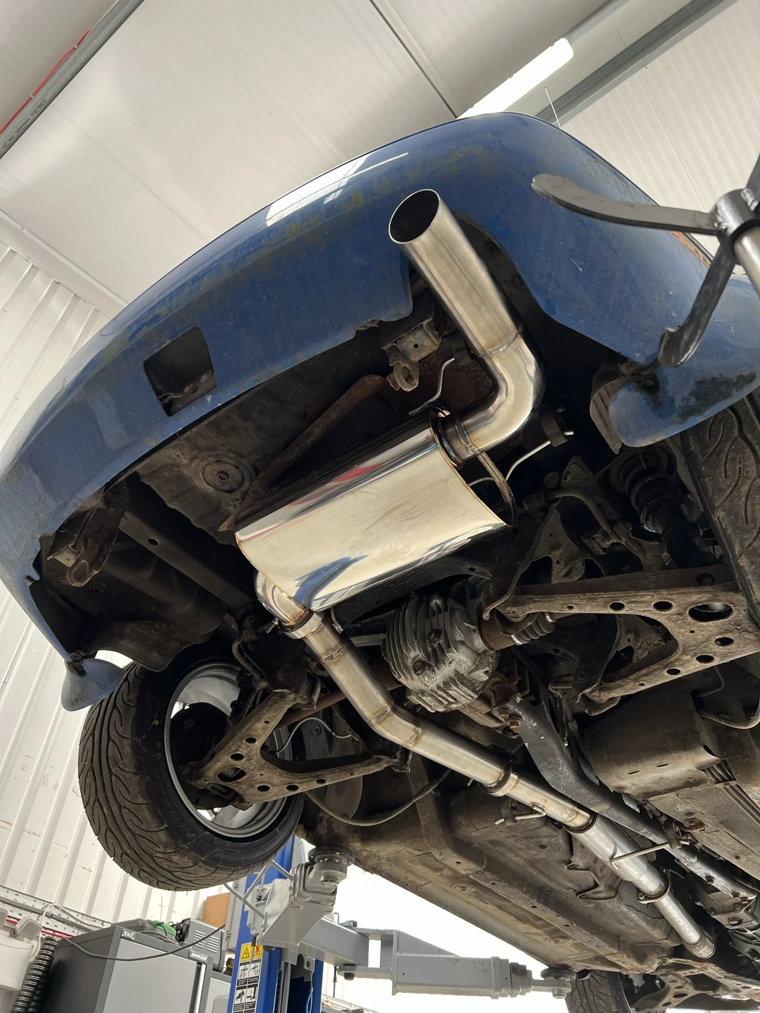 Underside of a blue car with a new chrome exhaust system installed, raised on a vehicle lift in a garage or workshop. Mazda Miata MX5 MK1 Eunos 