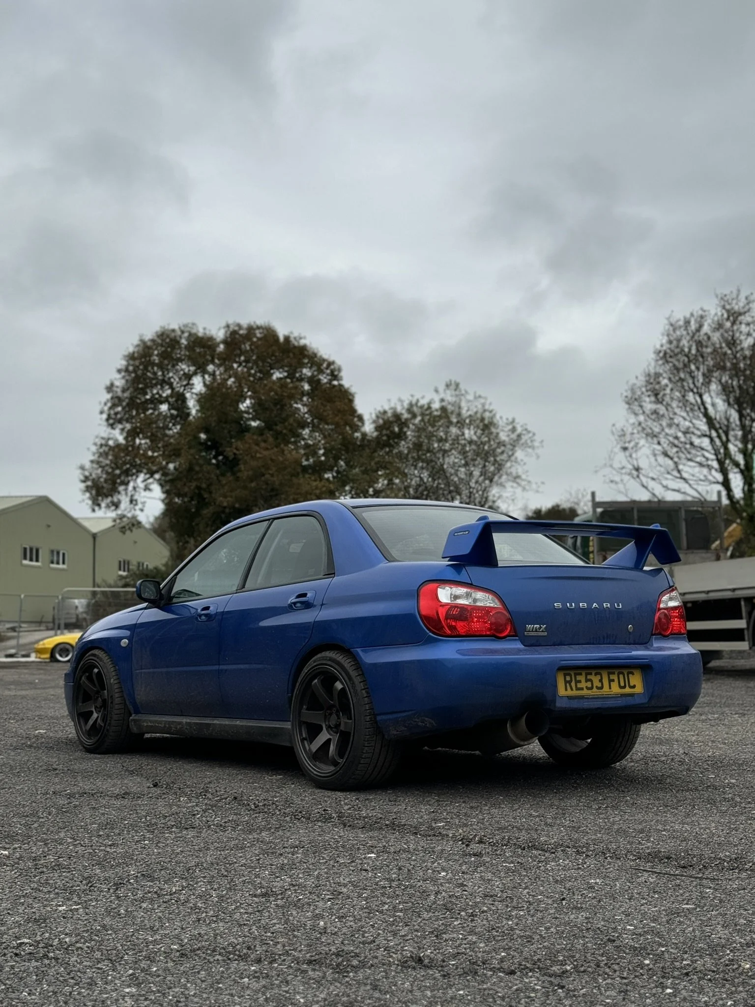 Blue Subaru WRX parked on gravel with trees and industrial buildings in the background, under cloudy sky.