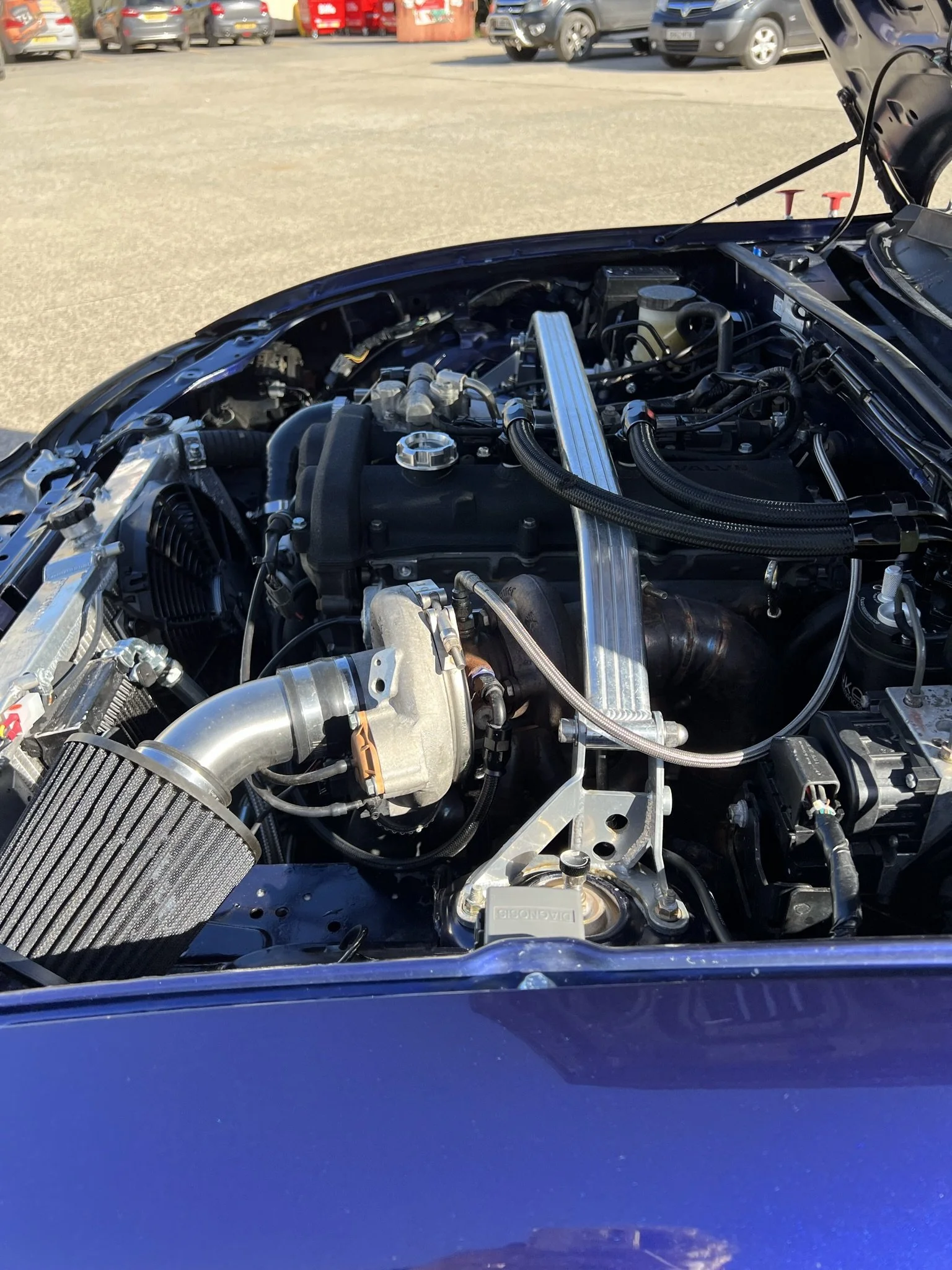 The image shows the engine bay of a mazda mx5, with various engine components including a large air filter, hoses, and metal parts, in a parking lot with other parked vehicles in the background.