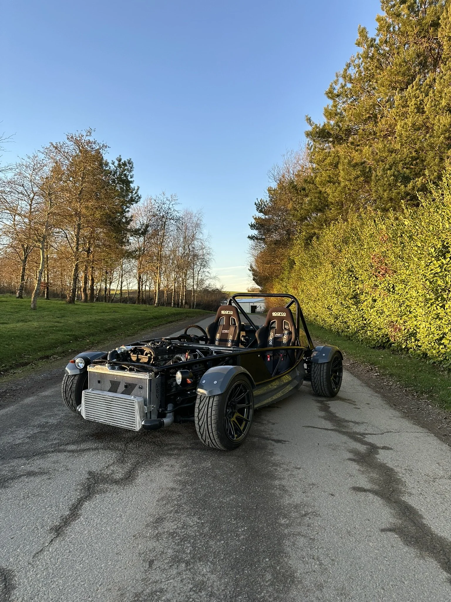 An open-frame, black race car with large tires and two seats parked on a rural road with trees on both sides and a clear blue sky. MEV Exocet