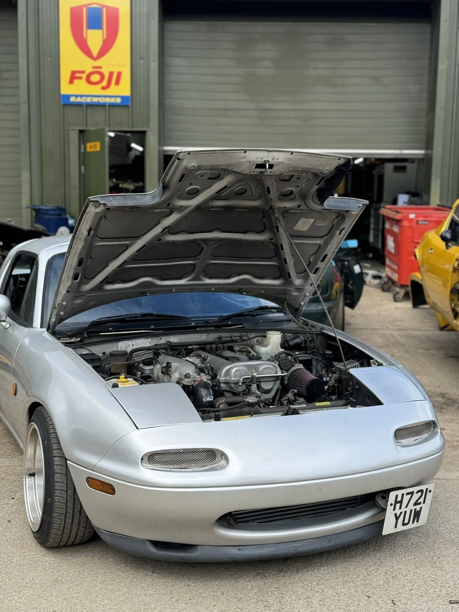 Mazda MX5 Miata Eunos Roadster. A silver sports car with its hood open, showing the engine, parked outside a raceway garage with a 'FOJI Raceworks' sign in the background.