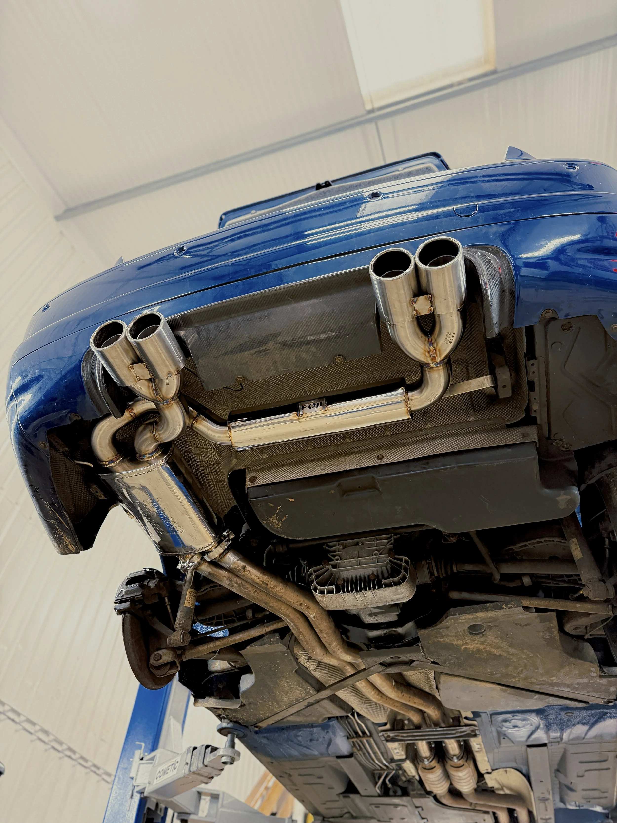 Underneath a blue sports car, showing modified exhaust pipes, a heat shield, and parts of the chassis. M3 E46 BMW