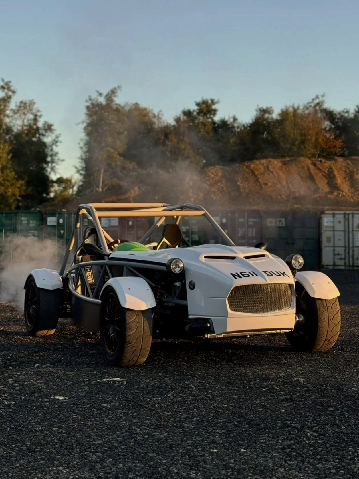 A white and black MEV exocet race car with a roll cage, parked on a gravel surface, with trees, a hillside, and shipping containers in the background, and dust clouds around the tires.