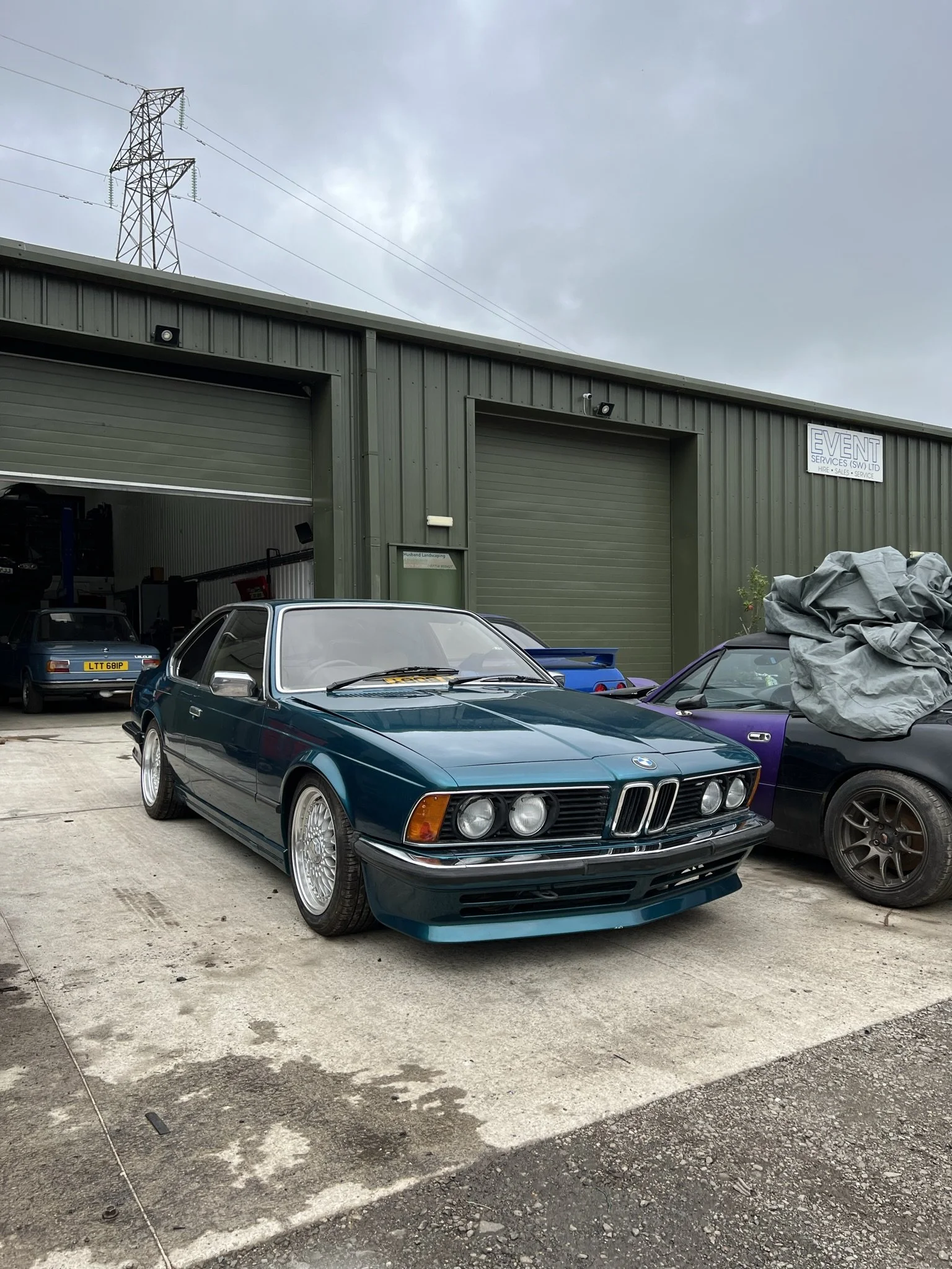 A teal-colored vintage BMW car parked outside a green industrial building, with other cars nearby, under an overcast sky.