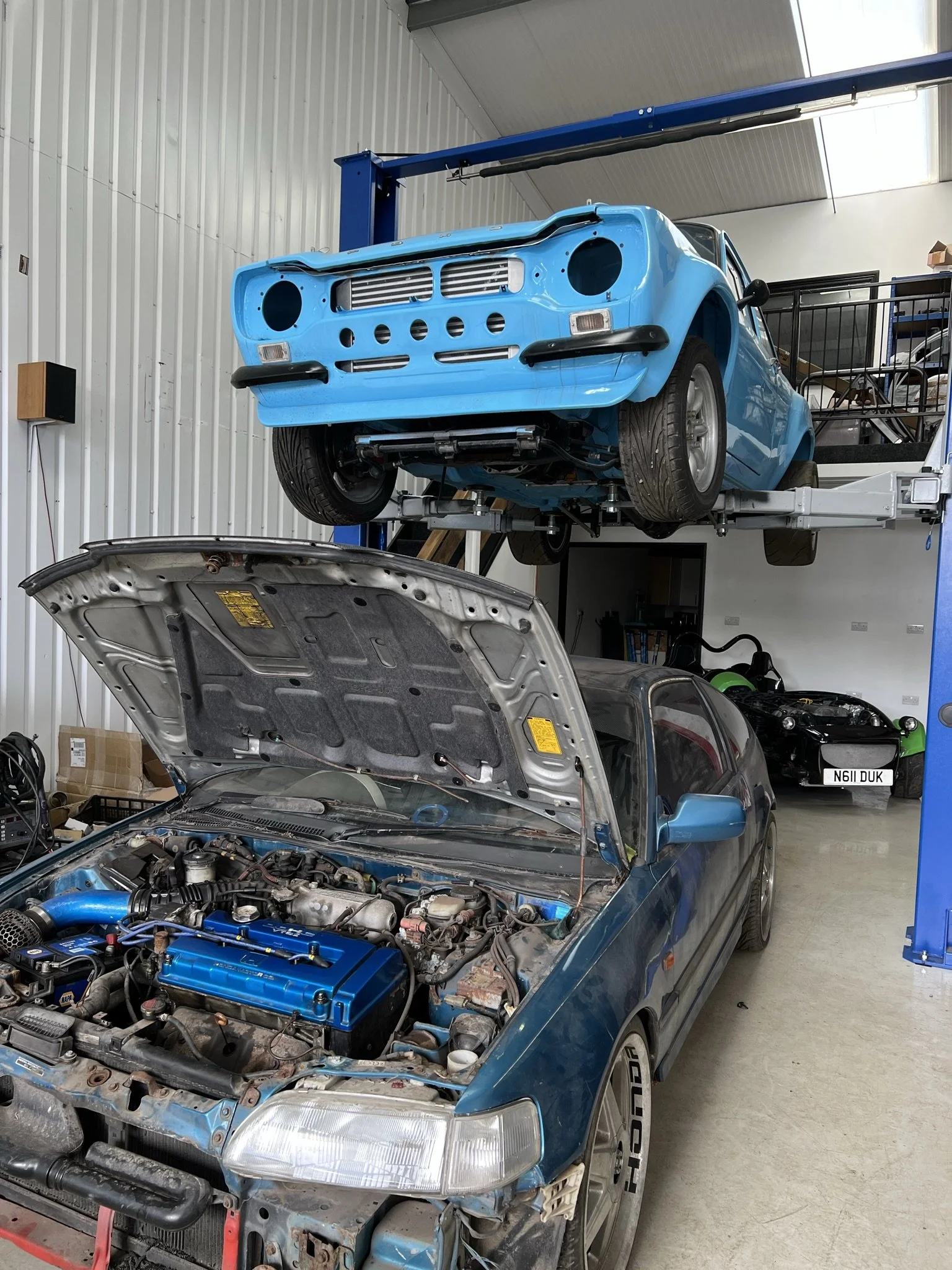 Two blue cars in a workshop, one with its hood open revealing the engine, the other elevated on a lift without a front bumper, in a garage with a collection of other vehicles and auto parts. HONDA CRX FORD ESCORT MK!
