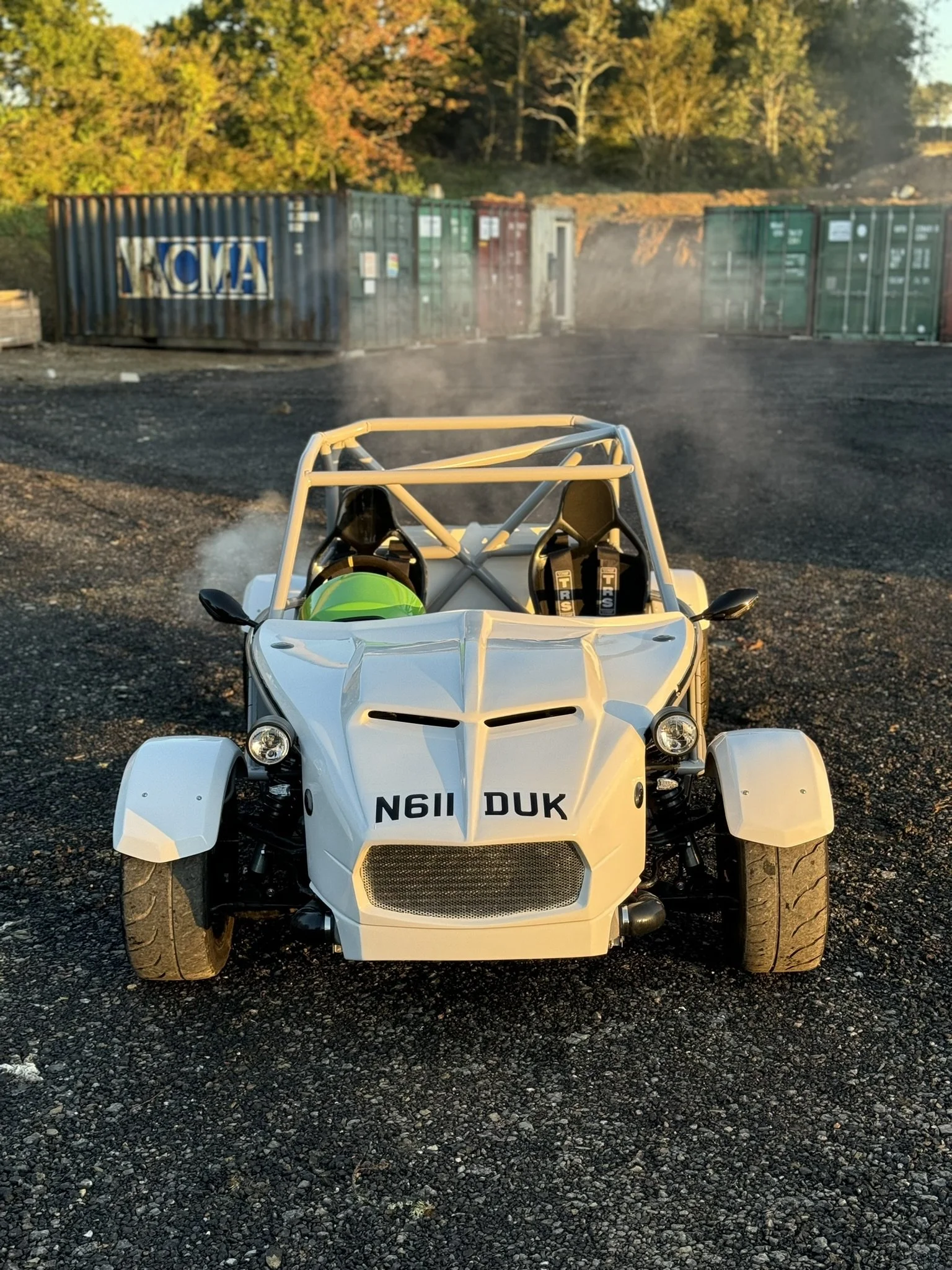 Front view of a white open-top race car with a roll cage, parked on a gravel lot during sunset. A green helmet is visible inside, and two racing seats with harnesses are present. MEV Exocet By Foji Raceworks
