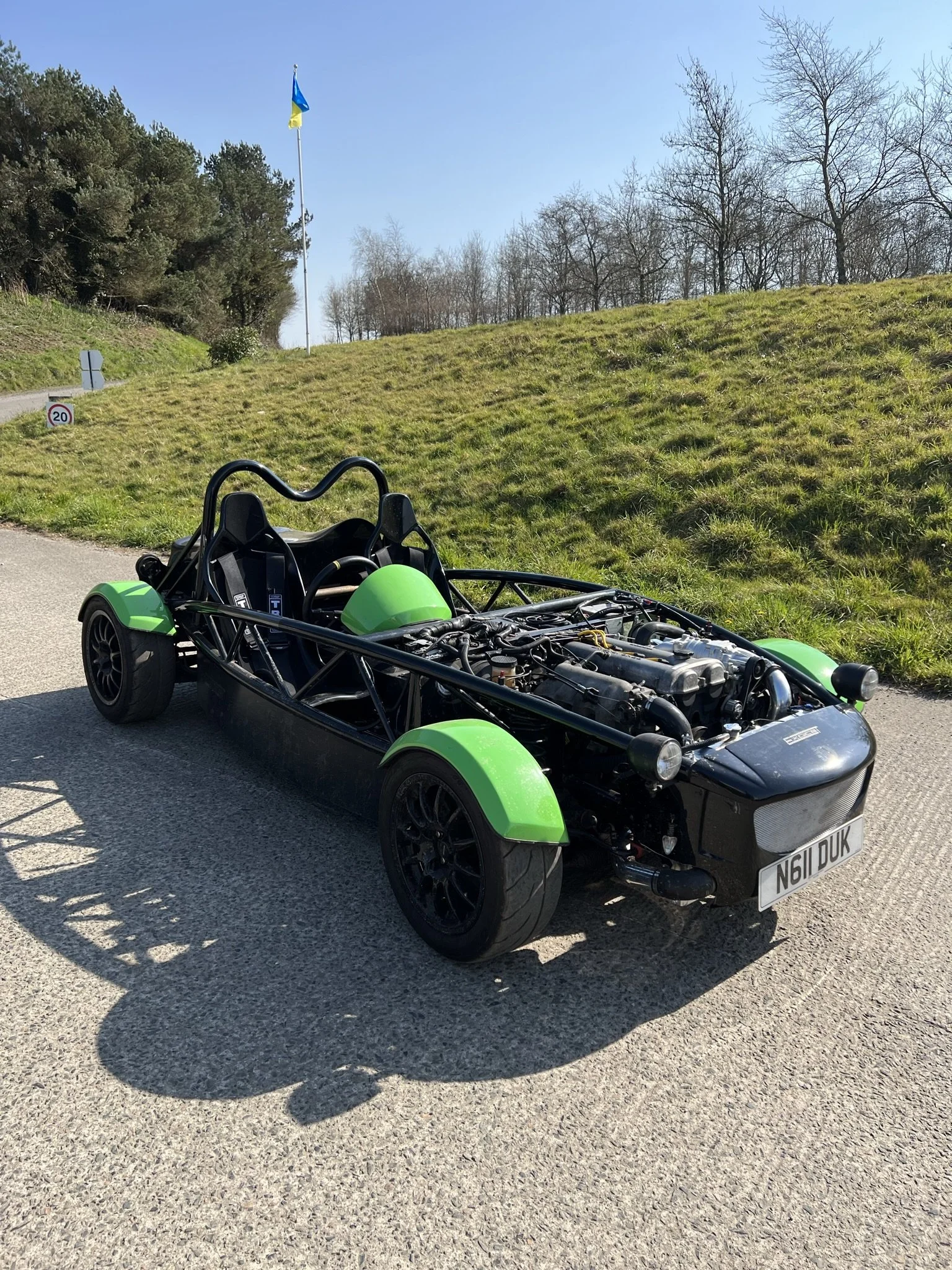 A green and black open-frame sports car with a visible engine, parked on a paved road near grass and trees, with a blue and yellow flag flying on a pole in the background. MEV Exocet