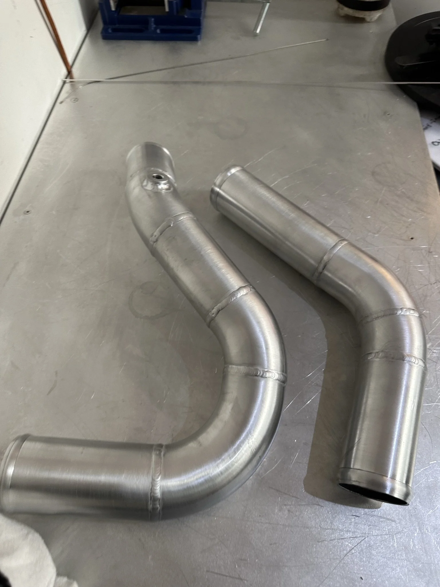 Two silver metal pipes with bends placed on a metal work surface in a workshop or garage.