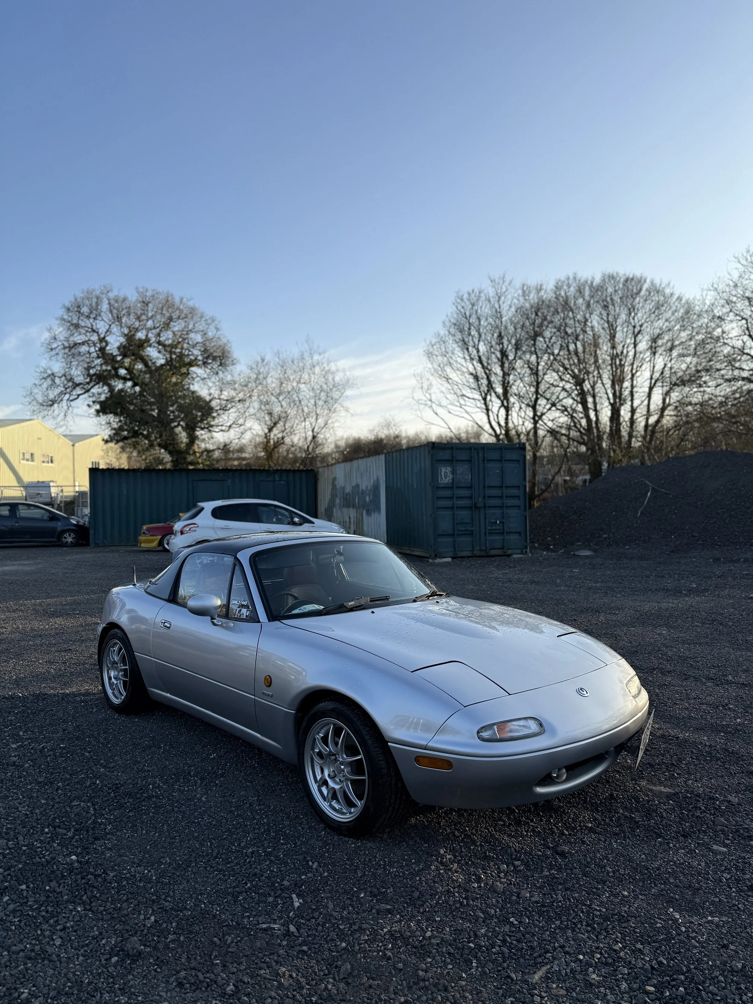 Silver Mazda MX-5 Miata convertible parked on gravel lot with trees, shipping containers, and other cars in the background under a clear blue sky.