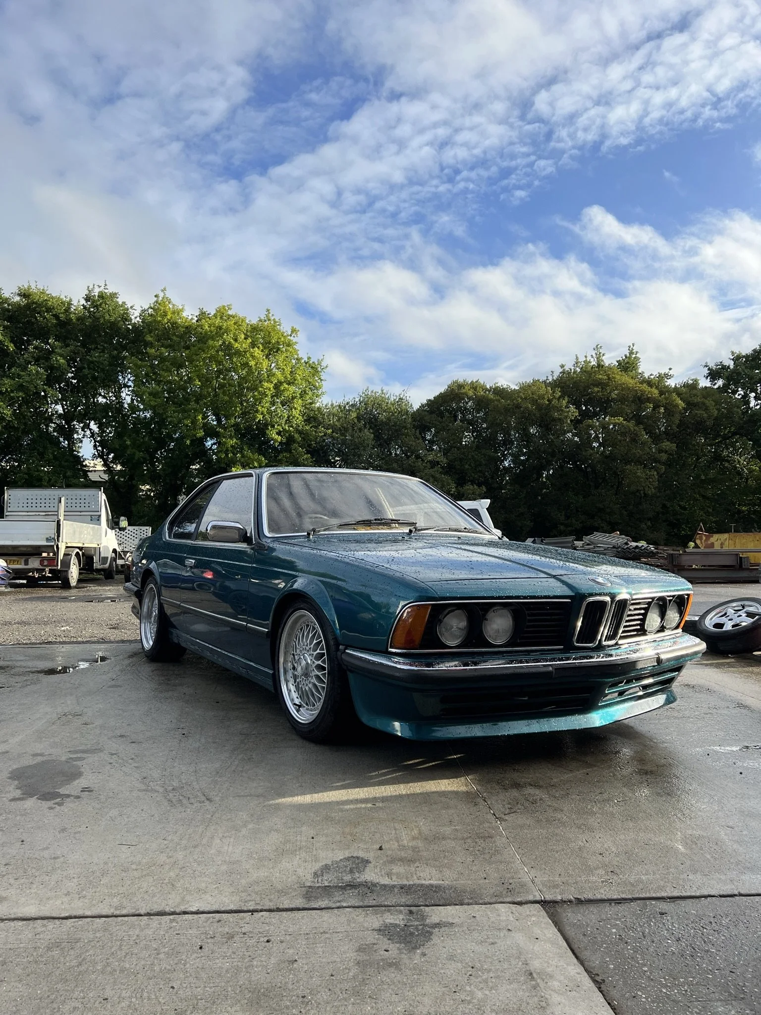 A vintage teal BMW E24 6 Series coupe parked on a concrete surface with puddles, surrounded by trees and other vehicles in the background, under a partly cloudy sky.