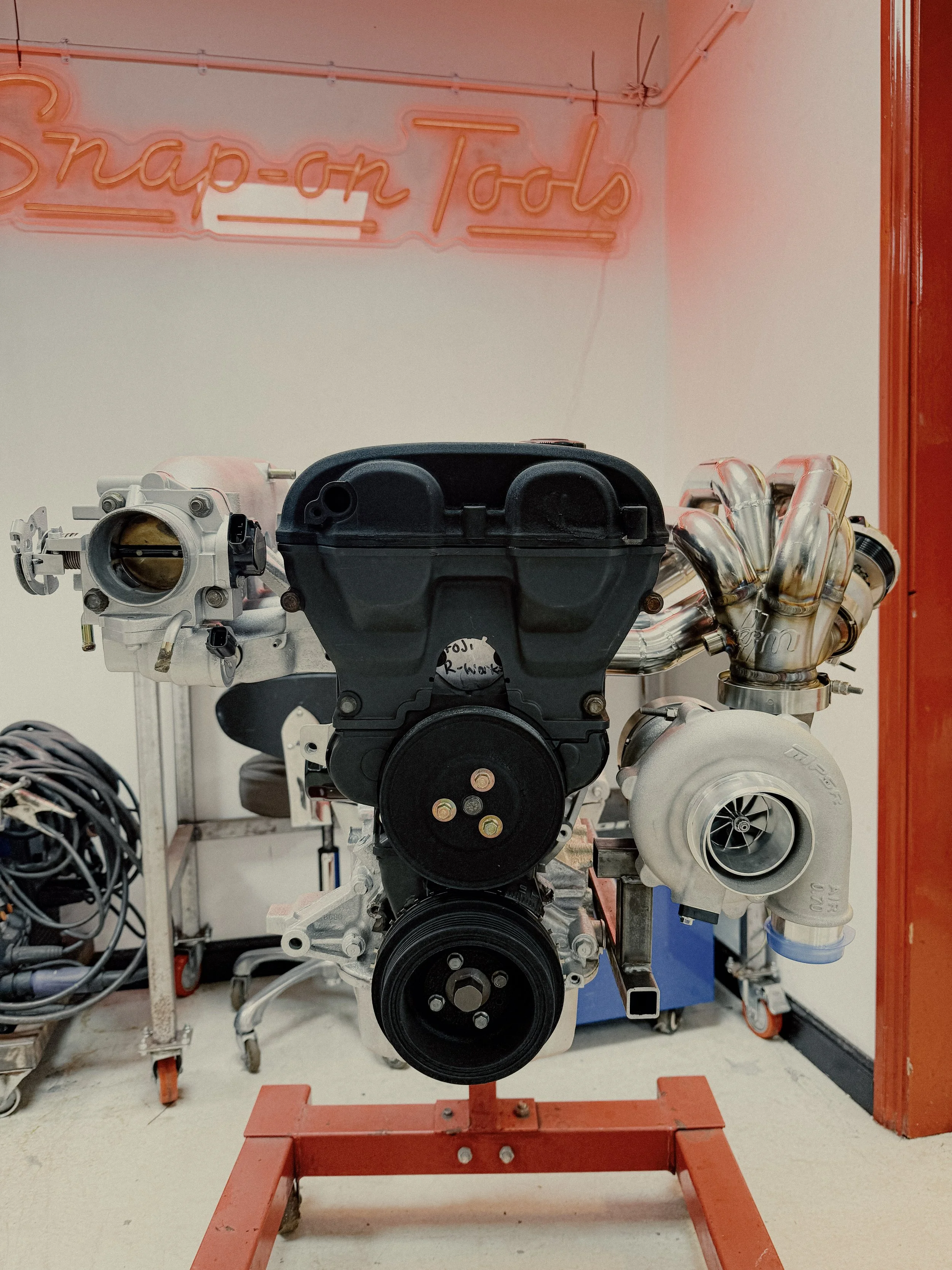 A car engine on a red stand in a workshop with a neon sign reading 'Snap-on Tools' in the background. Mazda Miata MX5 MK1 Eunos  Turbo Manifold