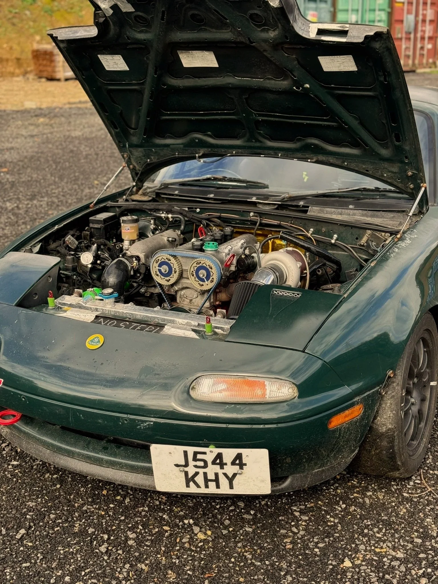 Green Mazda Mx5 Miata Mk1 sports car with the hood open, showing a modified engine with aftermarket parts, located on a gravel lot.