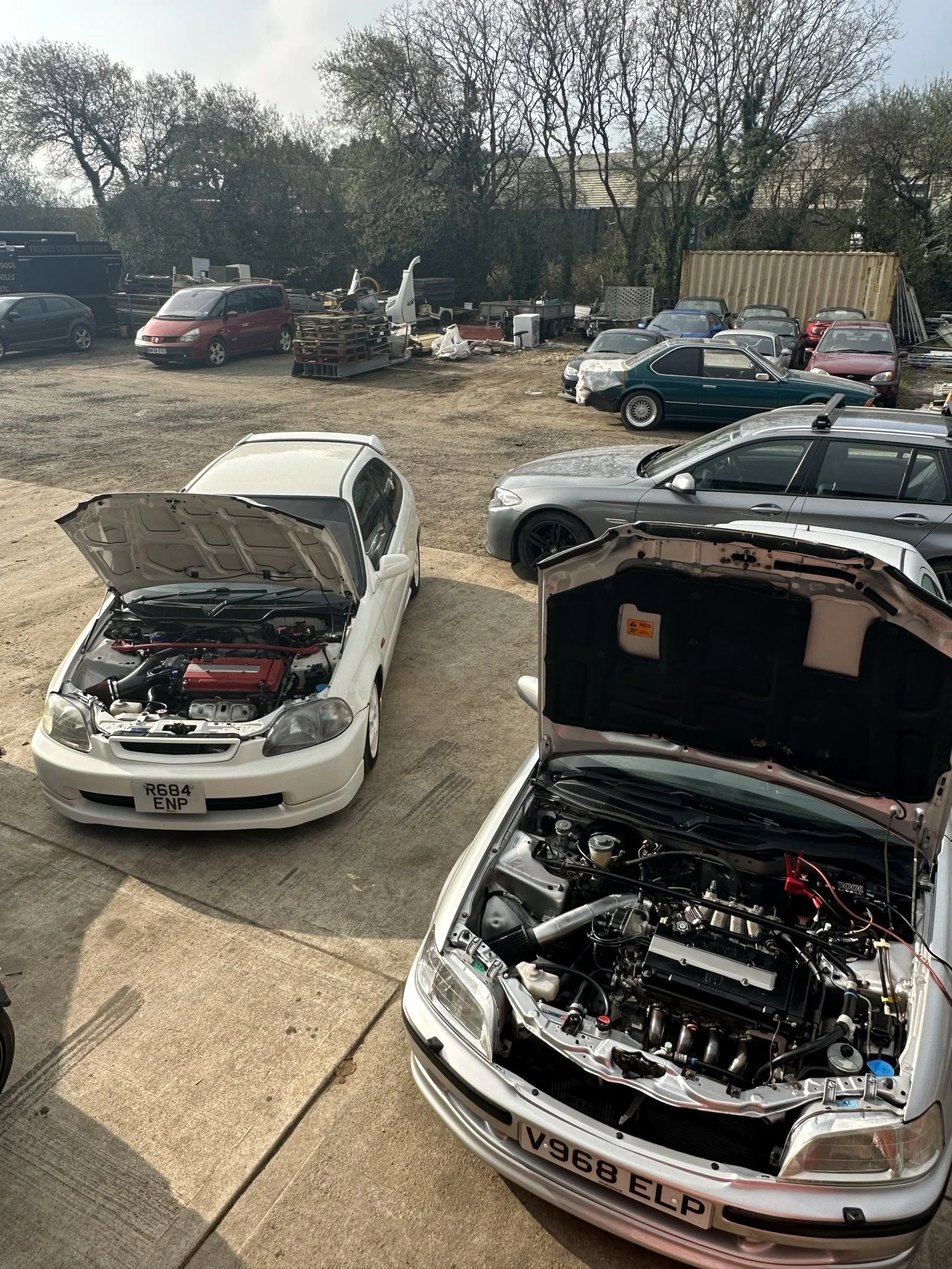 Two cars with hoods open parked in front of a gravel lot filled with various vehicles, some with partially removed or damaged parts, and a shipping container in the background, with trees and a residential area behind. HONDA CIVIC MB6 EK9