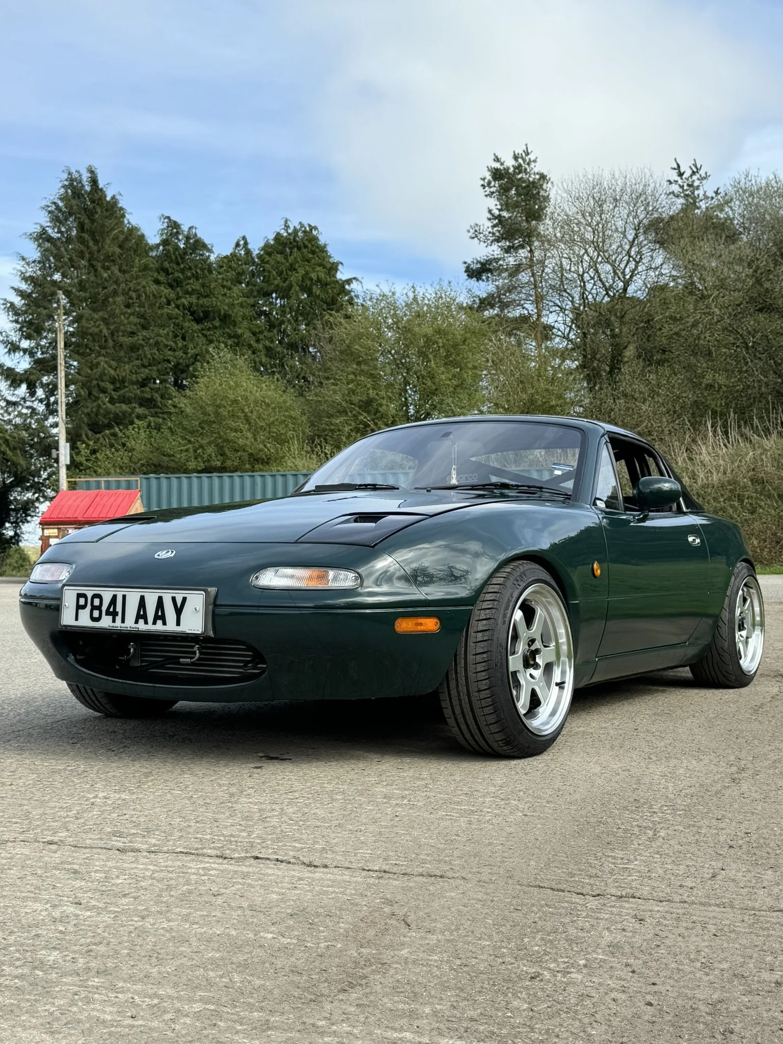 Green Mazda MX-5 Miata sports car parked on a paved surface with trees and a cloudy sky in the background.