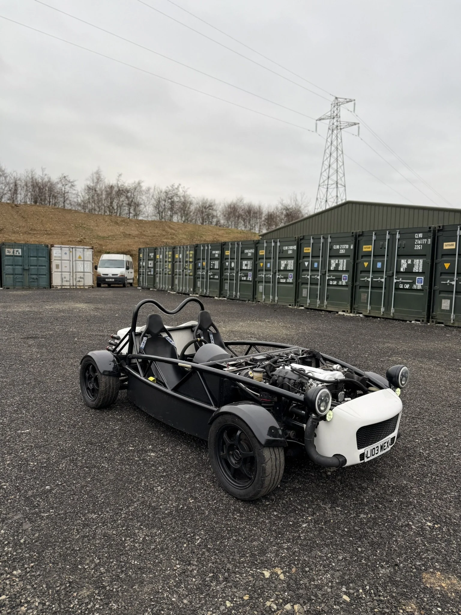 A partially assembled black and white open-frame race car or kit car parked on a gravel surface with green storage containers and a white van in the background, and electrical transmission towers overhead. MEV Exocet