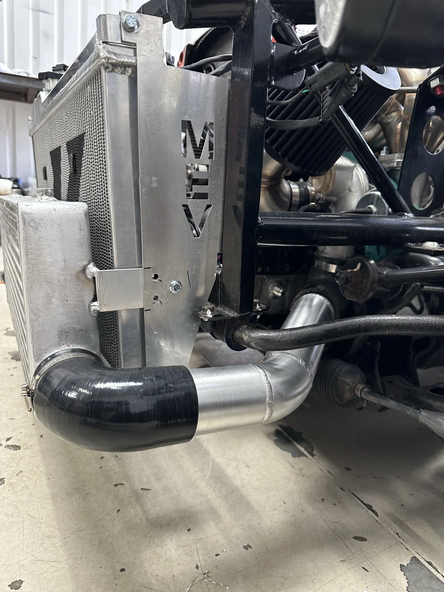 Close-up of a vehicle's cooling system and engine components, featuring a large radiator with a metallic finish and a cutout with the brand name 'MEV,' along with a black and silver pipe.