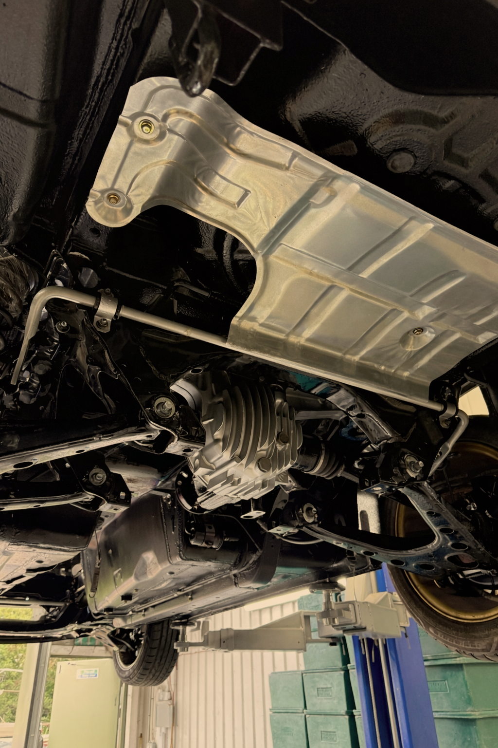 Underbody view of a car showing a large silver heat shield, engine components, and suspension parts in a garage or workshop. The underbody of a mazda mx5