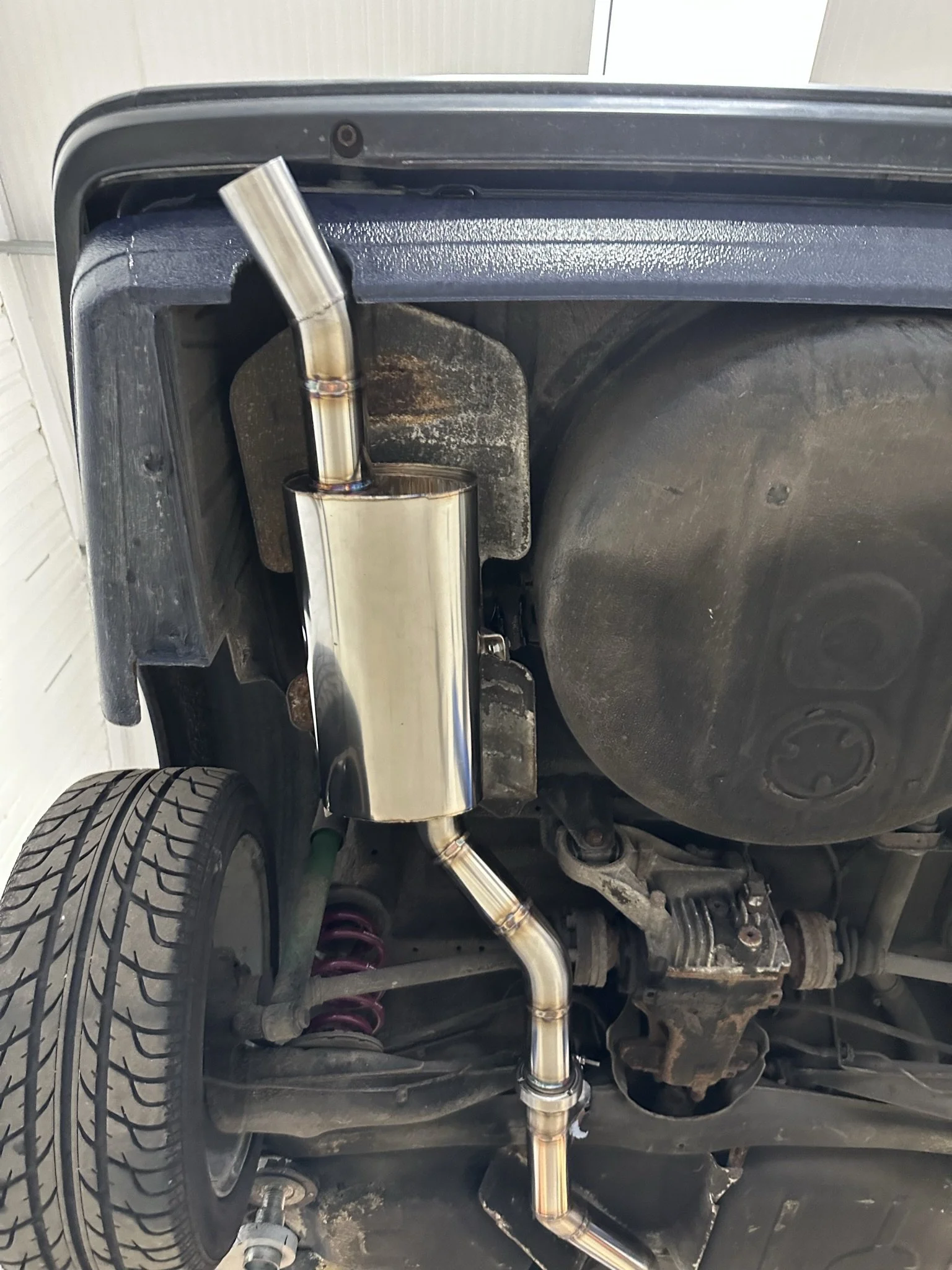 Underneath a vehicle showing a new stainless steel exhaust system with pipes and muffler, along with parts of the suspension and rear wheel. E30 BMW