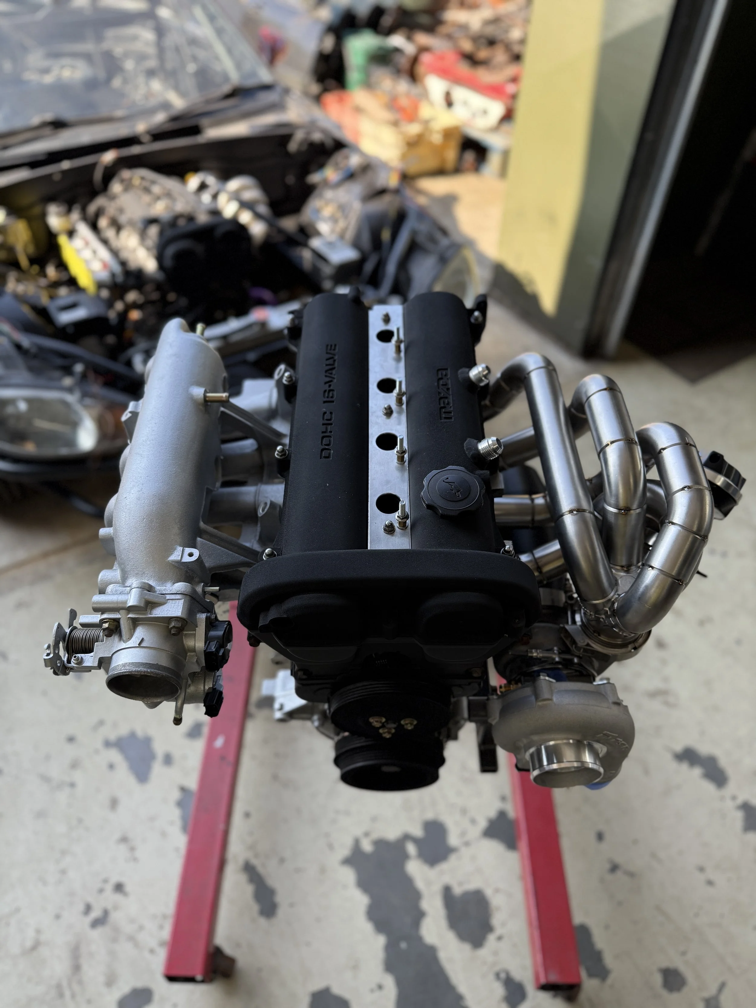 A car engine mounted on a red stand in a garage or workshop with various car parts and tools in the background. Turbo Mazda MX5 Miata 