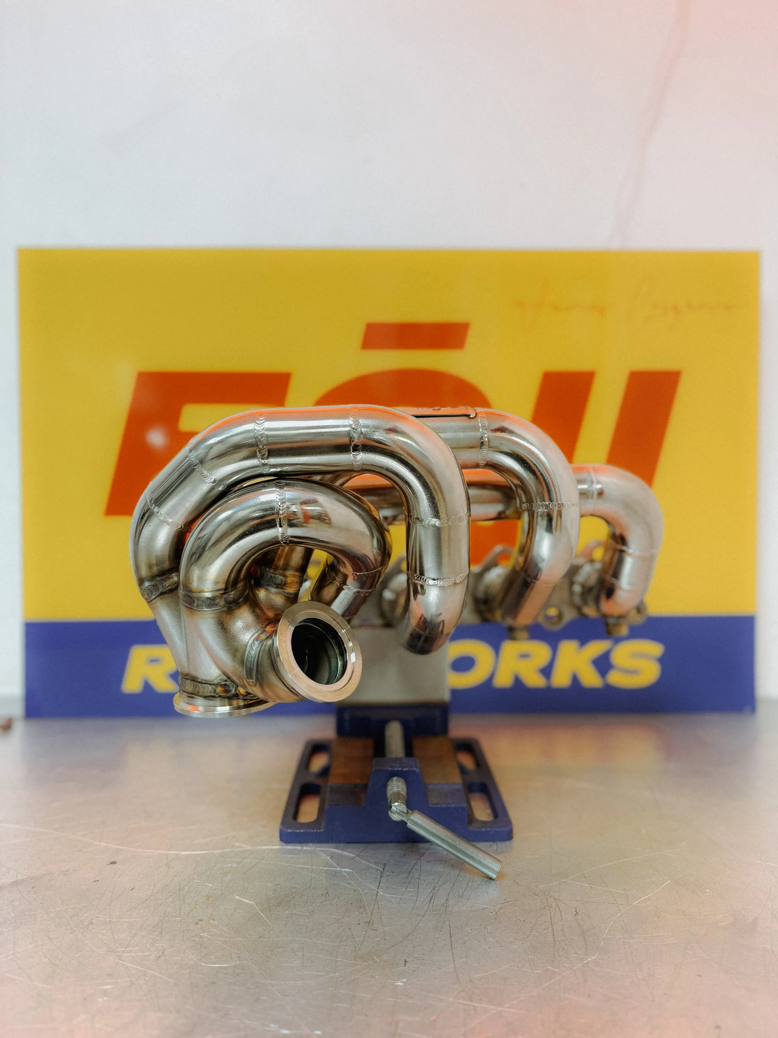 A metal exhaust manifold for an automotive engine, mounted on a blue stand, with a yellow and blue box labeled 'FOJI RACEWORKS' in the background. Mazda Miata MX5 MK1 Eunos turbo manifold