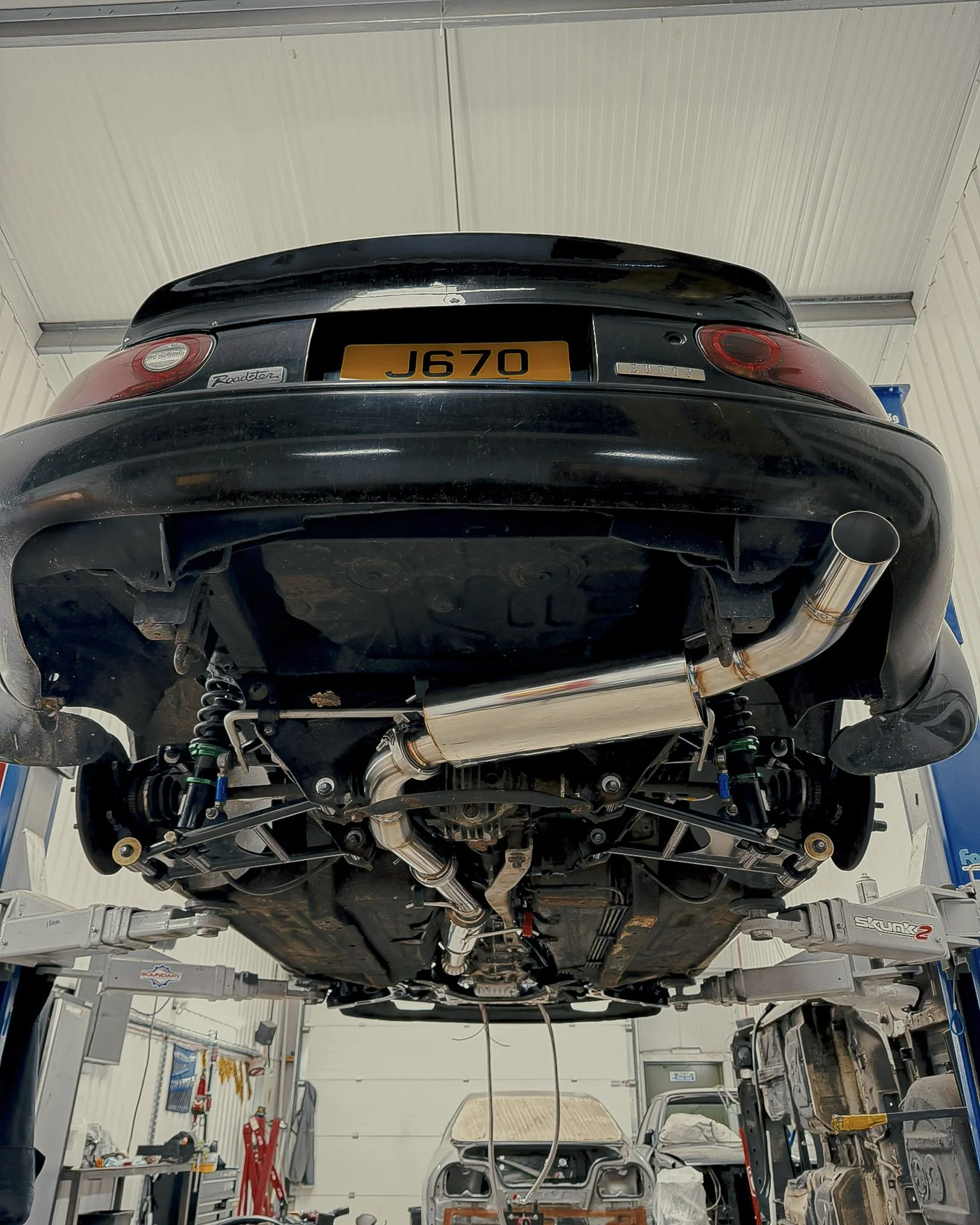 Underneath view of a black Mazda Miata MX5 MK1 Eunos  car on a lift showing the exhaust system and suspension components in a garage.