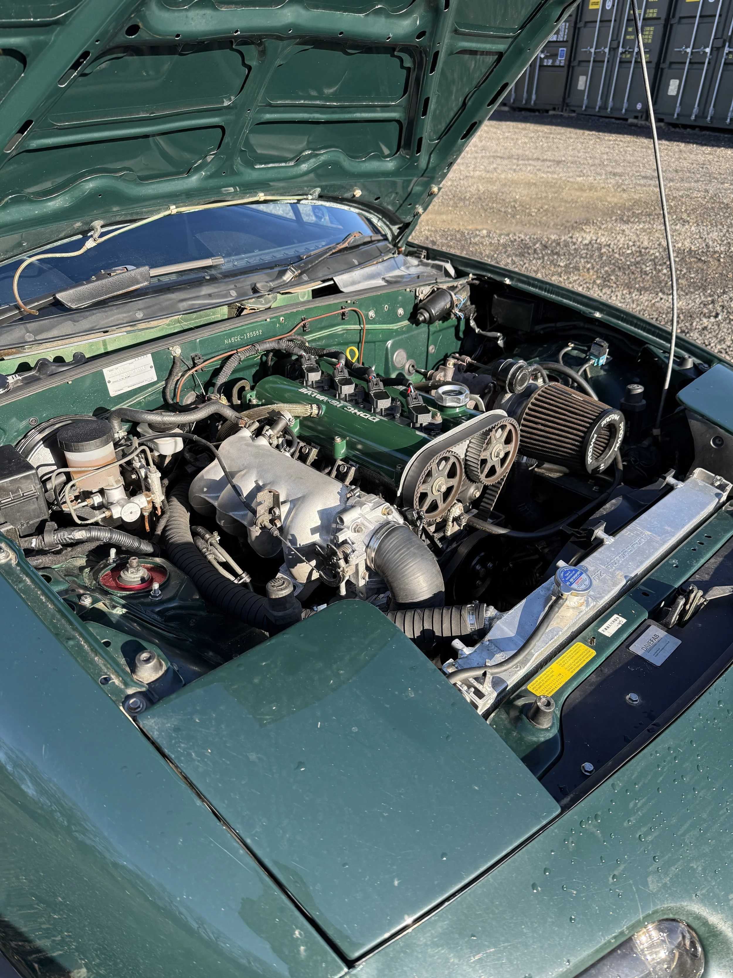 Green sports car with its hood open revealing a clean engine compartment with aftermarket parts, belts, and filters. Mazda mx5 miata mk1
