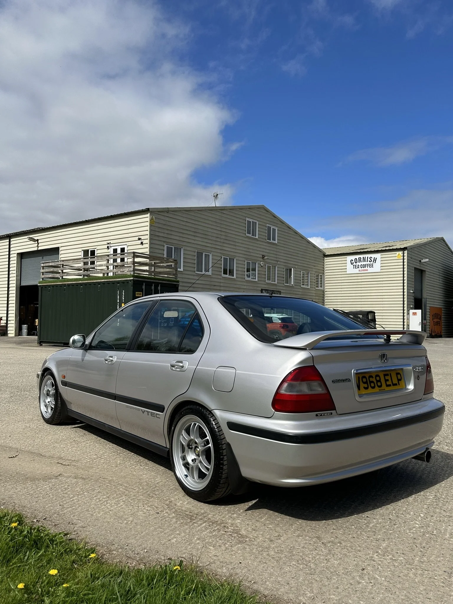 A silver Honda Civic hatchback with a spoiler parked on a concrete surface in front of industrial buildings under a partly cloudy sky.