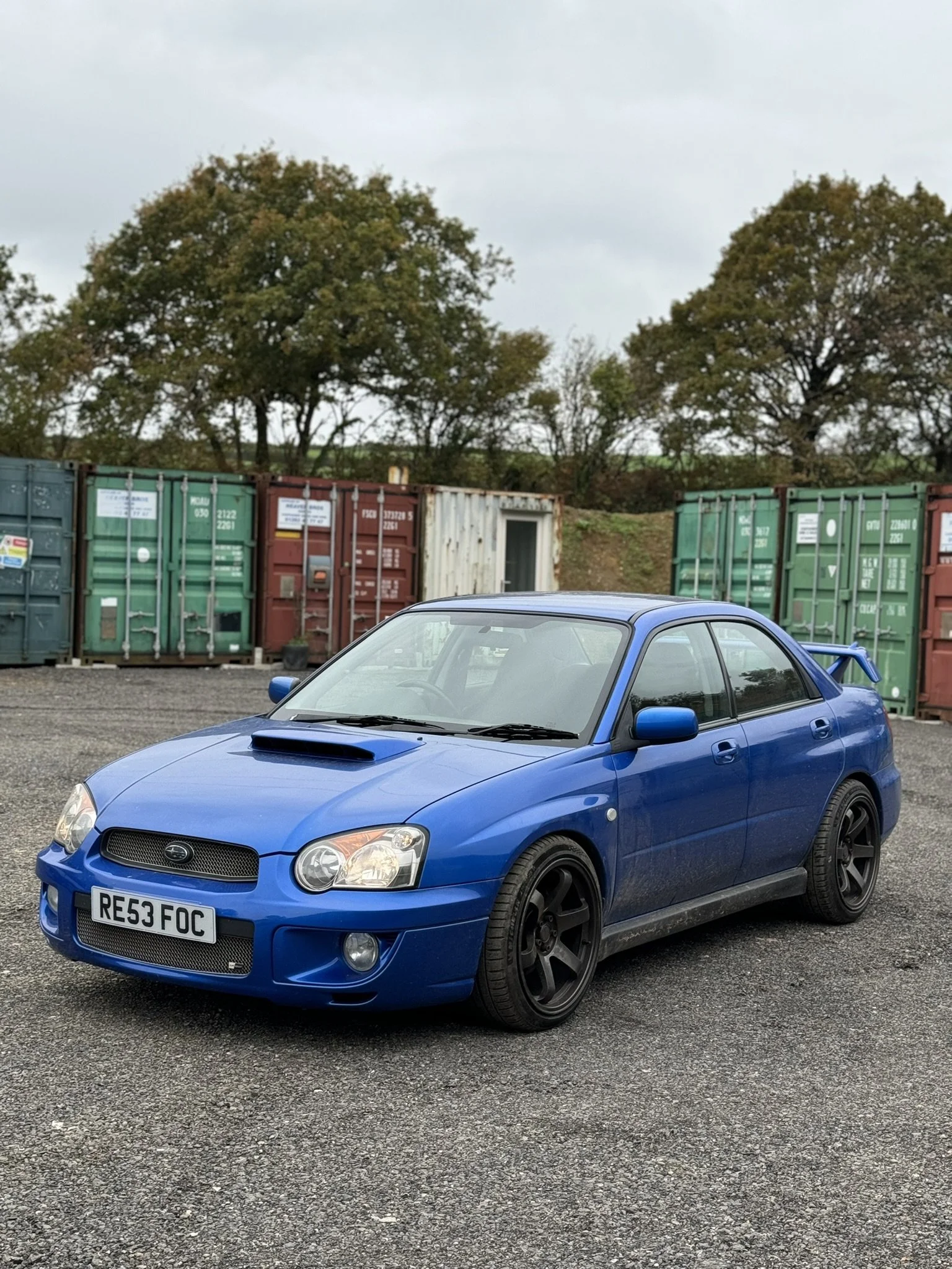 Blue Subaru Impreza parked on gravel with shipping containers and trees in the background.