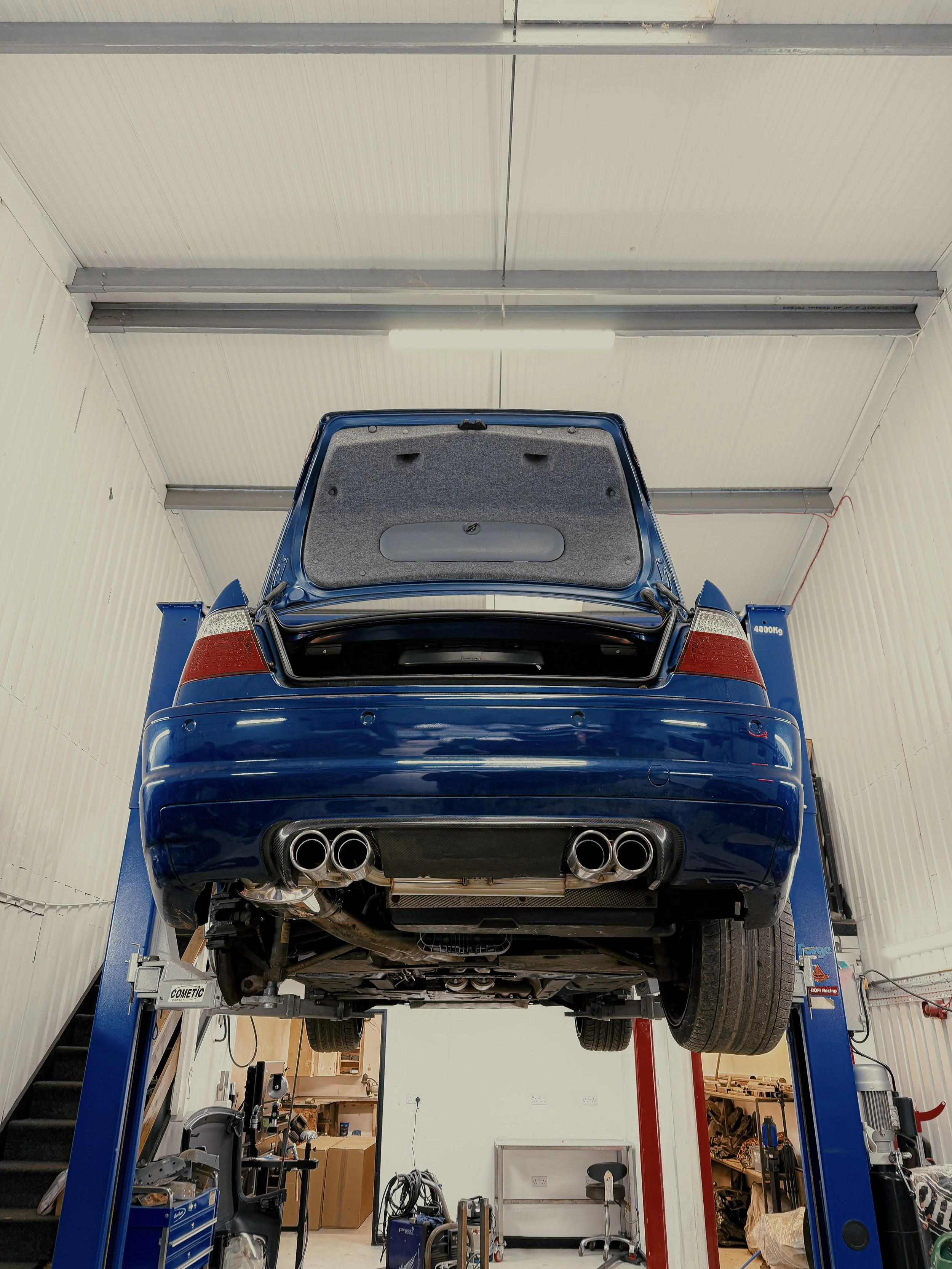 A blue sports car lifted on a hydraulic lift in an auto repair shop, showing the underside and exhaust system. E46 BMW M3