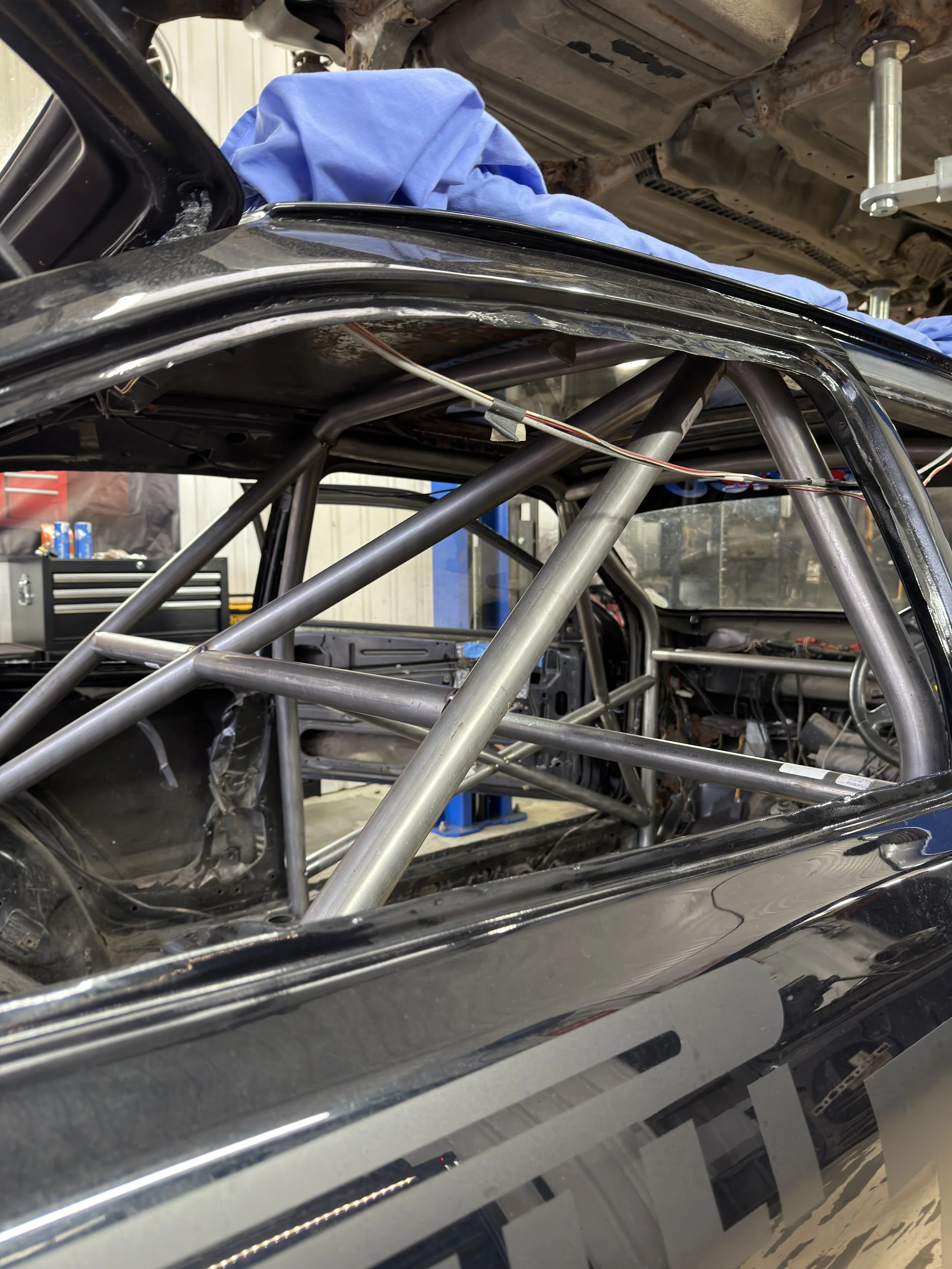 Inside a race car under construction, with a roll cage made of metal pipes visible inside the black body, and a blue cover draped over the exterior. FORD SEIRRA ROLL CAGE 
