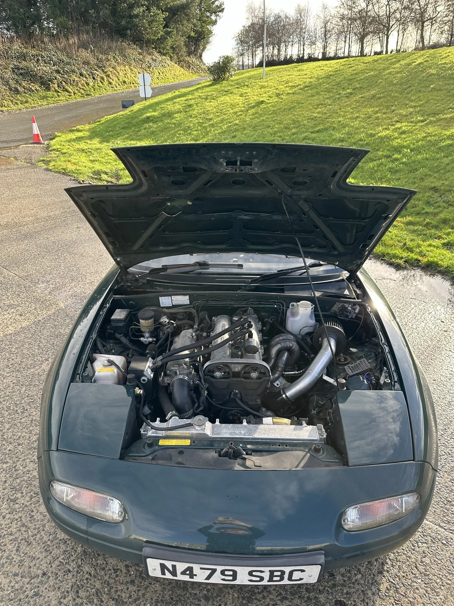 Front view of a dark green Mk1 mazda mx5 sports car with the hood open, showing the engine bay.
