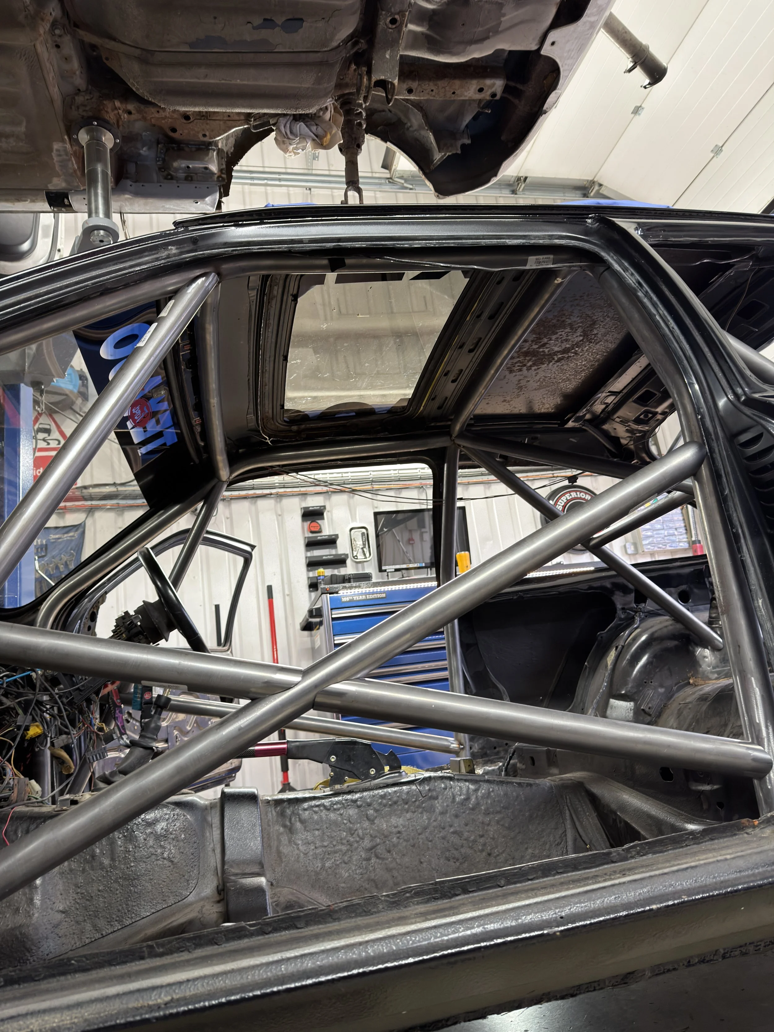 Inside a race car chassis with a roll cage, steering wheel, and dashboard removed, in a garage with tools and equipment visible in the background. FORD SEIRRA ROLL CAGE
