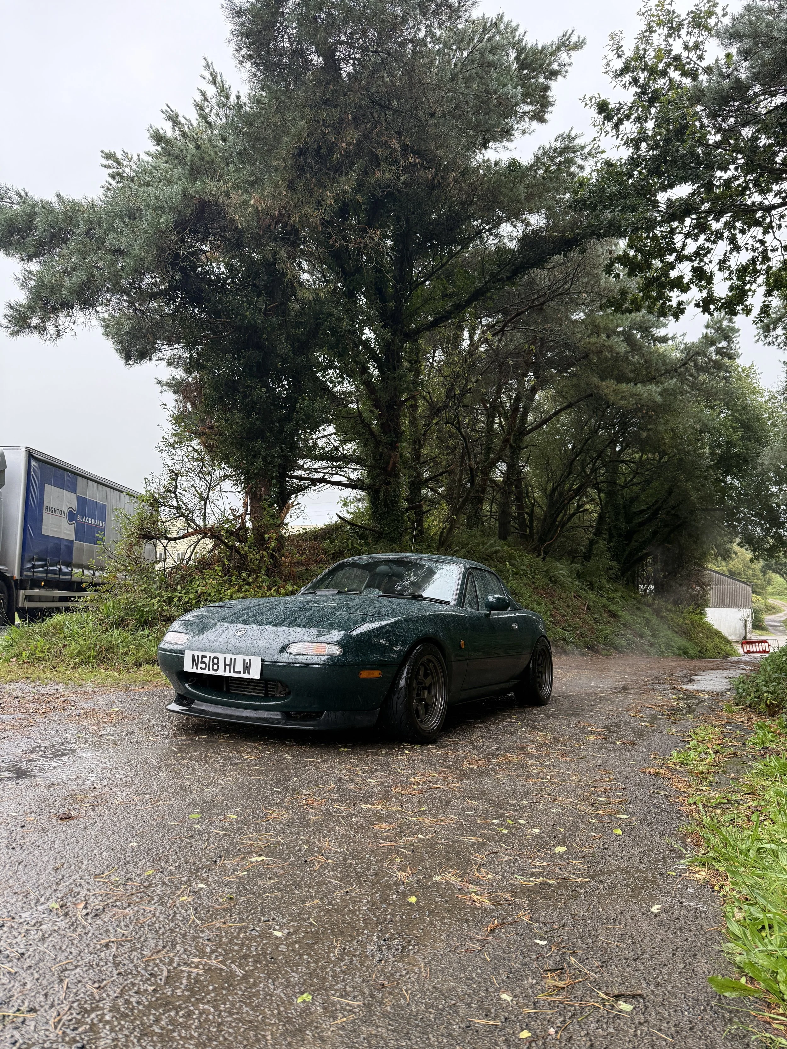 A dark green Mazda MX-5 Miata parked on a wet, gravel road near trees and bushes, with cloudy sky overhead on a rainy day.