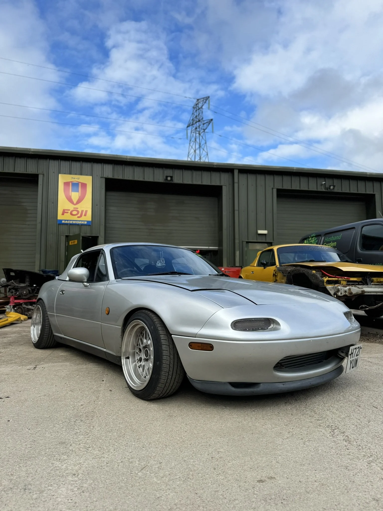 Silver Mazda Miata car parked outside at a raceway with a green building in the background, featuring a Foji Racewayworks sign, and a yellow sports car in the background.