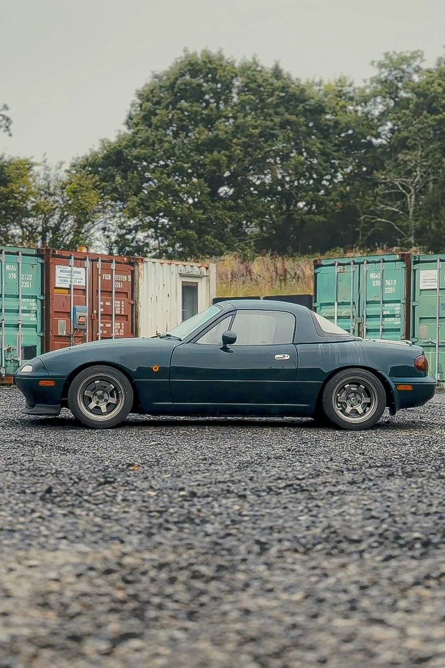 mazda mx5 mk1 eunos A classic black convertible sports car parked on gravel with colorful shipping containers and green trees in the background.