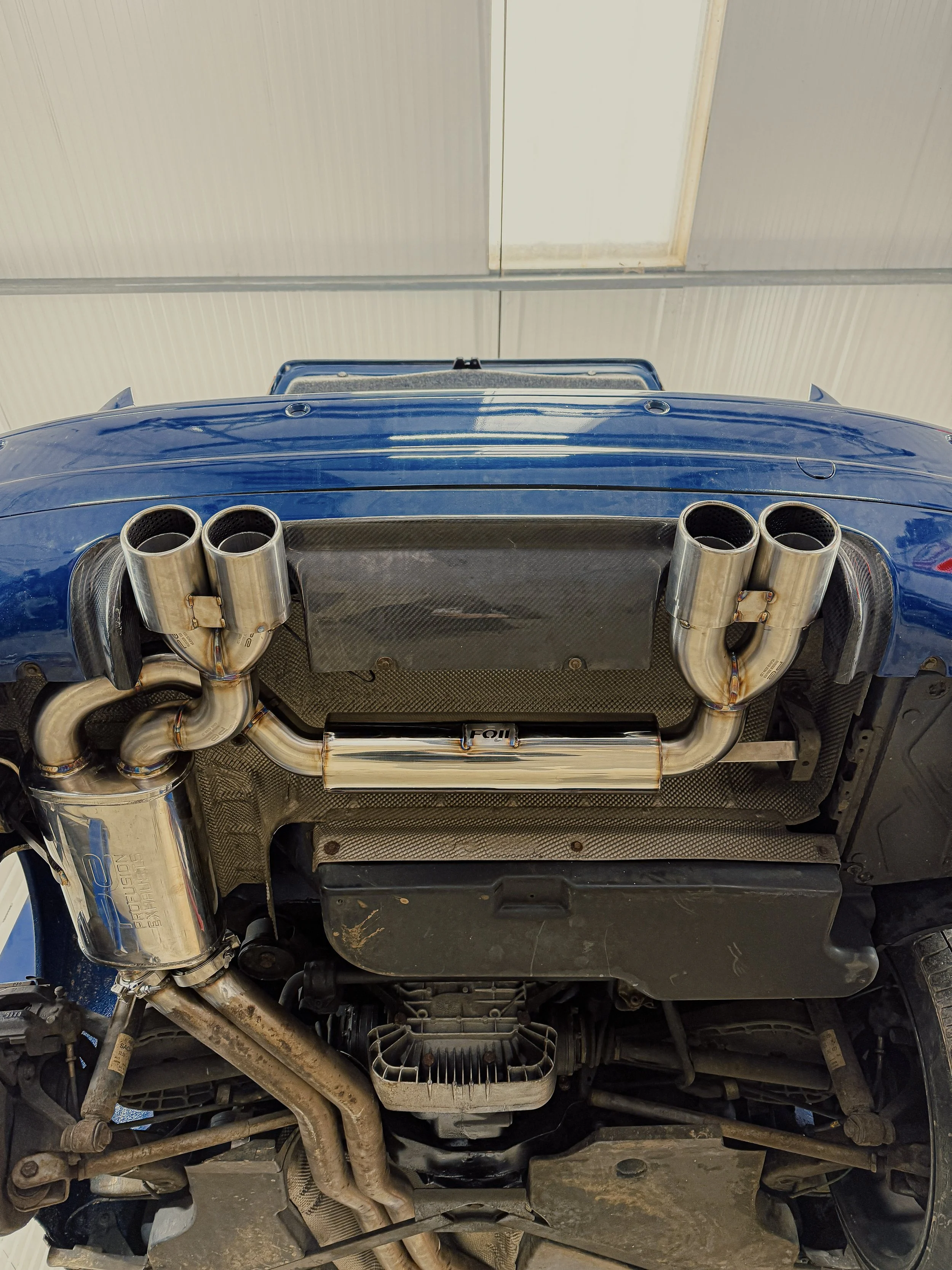 View of a blue car's underside showing exhaust system with twin pipes and catalytic converter, mounted on a lift inside a garage. E46 M3 BMW