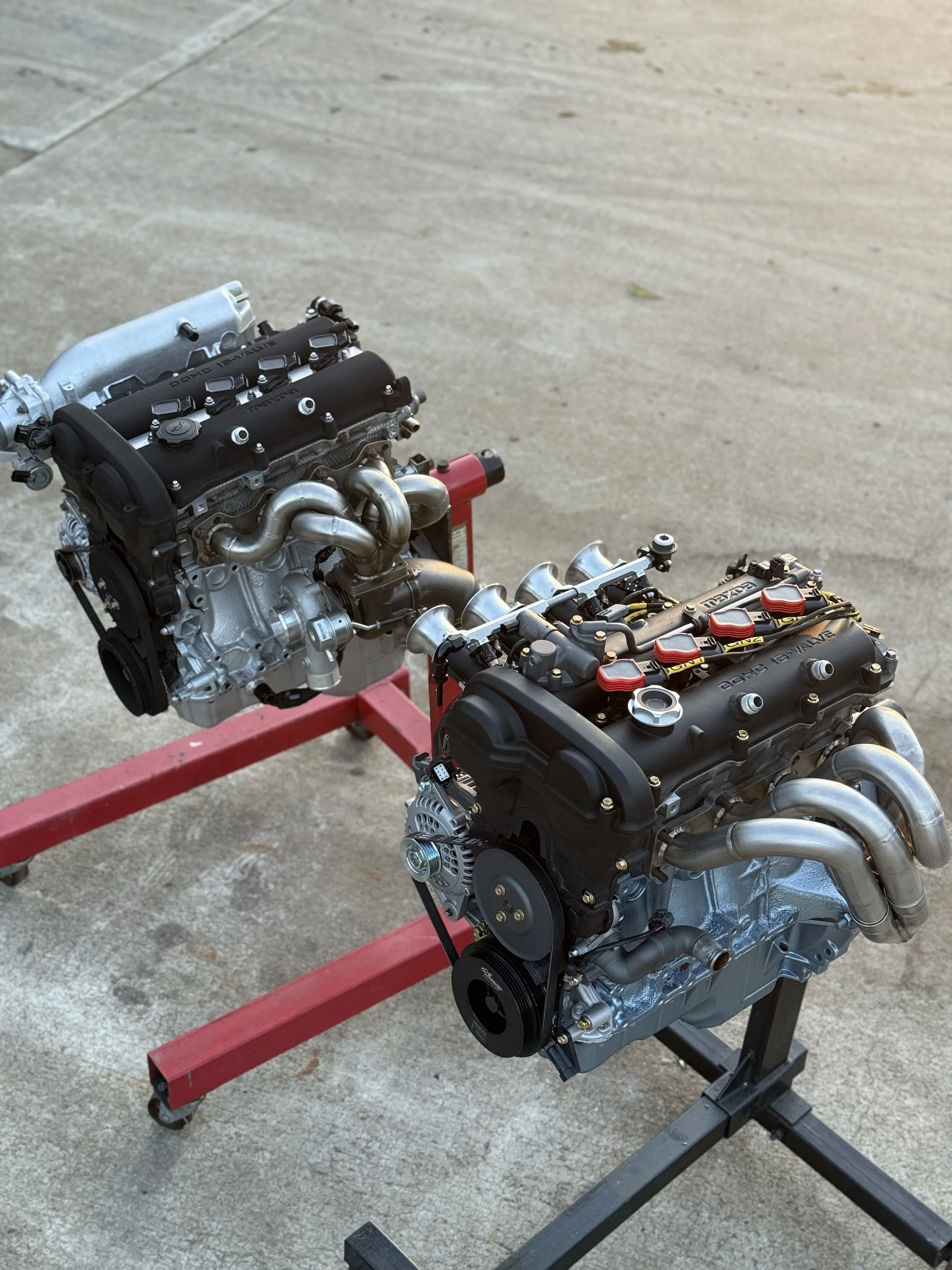 Two engine blocks mounted on a red stand outdoors on a concrete surface. Turbo mazda mx5 miata and a naturally aspirated version