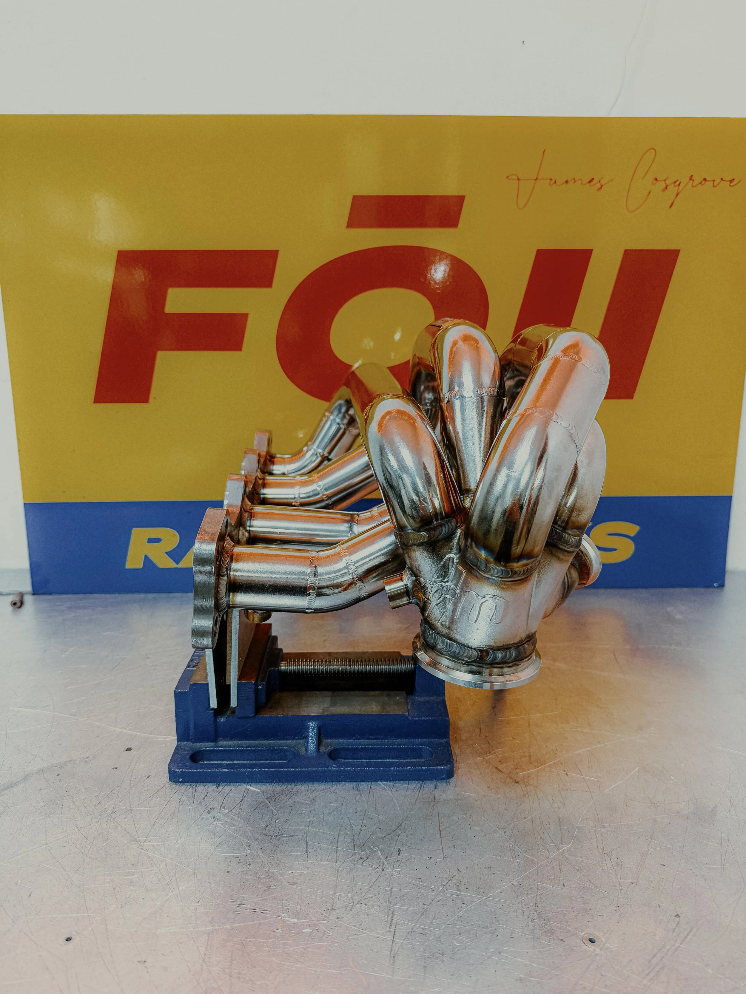 Small metallic sculpture of a car engine manifold mounted on a blue clamp, placed on a metallic surface with a yellow box behind it that has red and blue text. Mazda Miata MX5 MK1 Eunos  Turbo Manifold