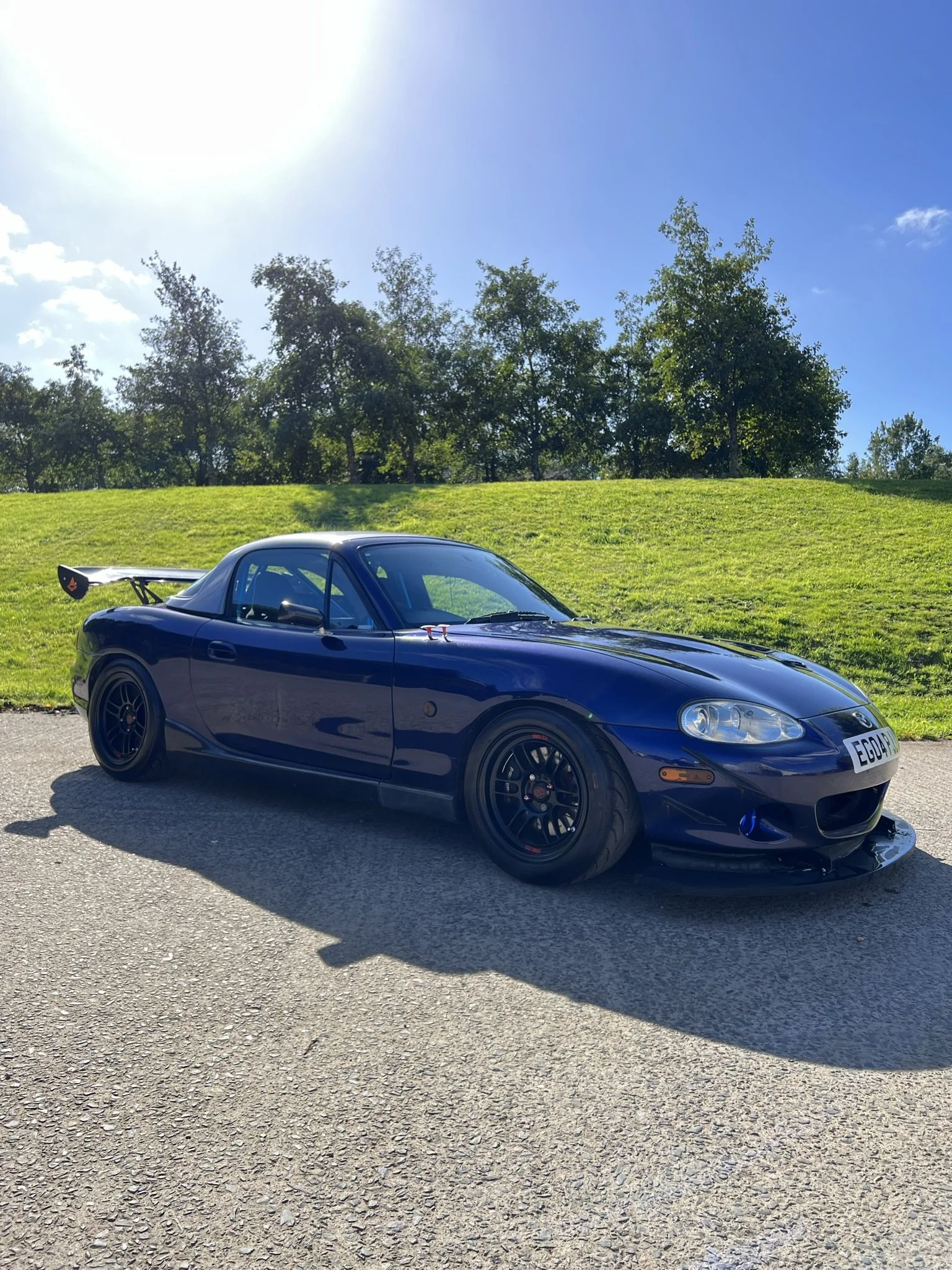 Mazda Mx5 miata A dark blue sports car with a rear wing, black wheels, and a sleek design parked on a road under a bright sun with a grassy hill and trees in the background.