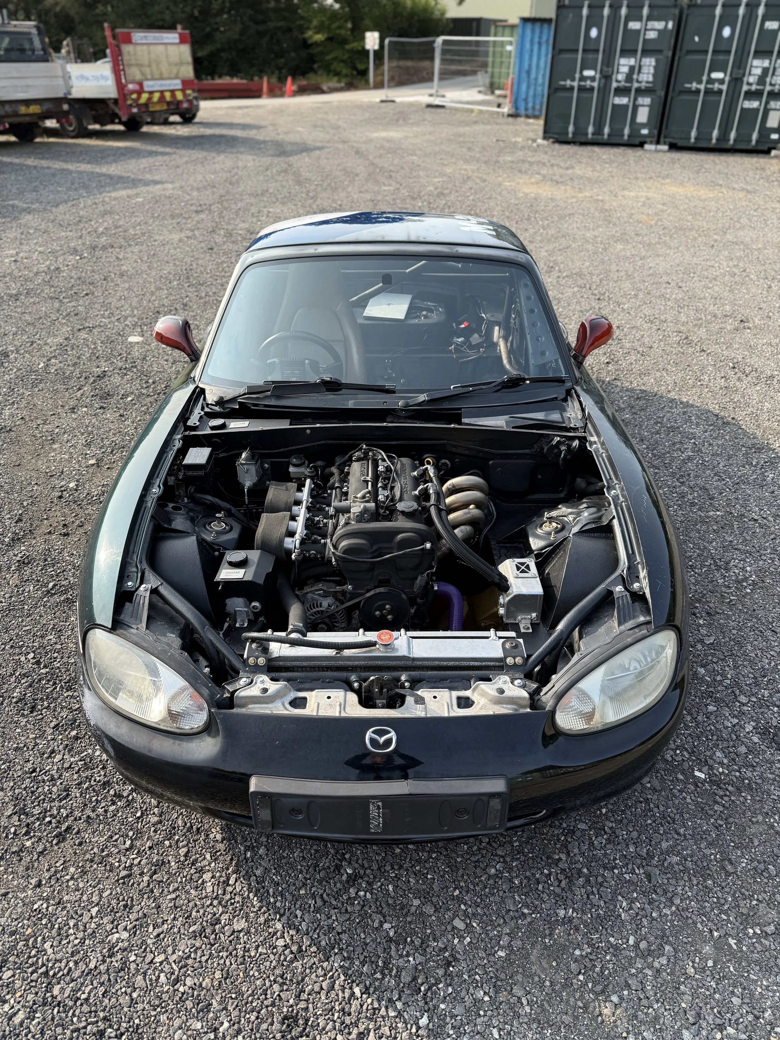 A black Mazda MX5 Miata sports car with its hood open, displaying the engine in an outdoor parking lot surrounded by gravel, nearby trucks, shipping containers, and fencing.