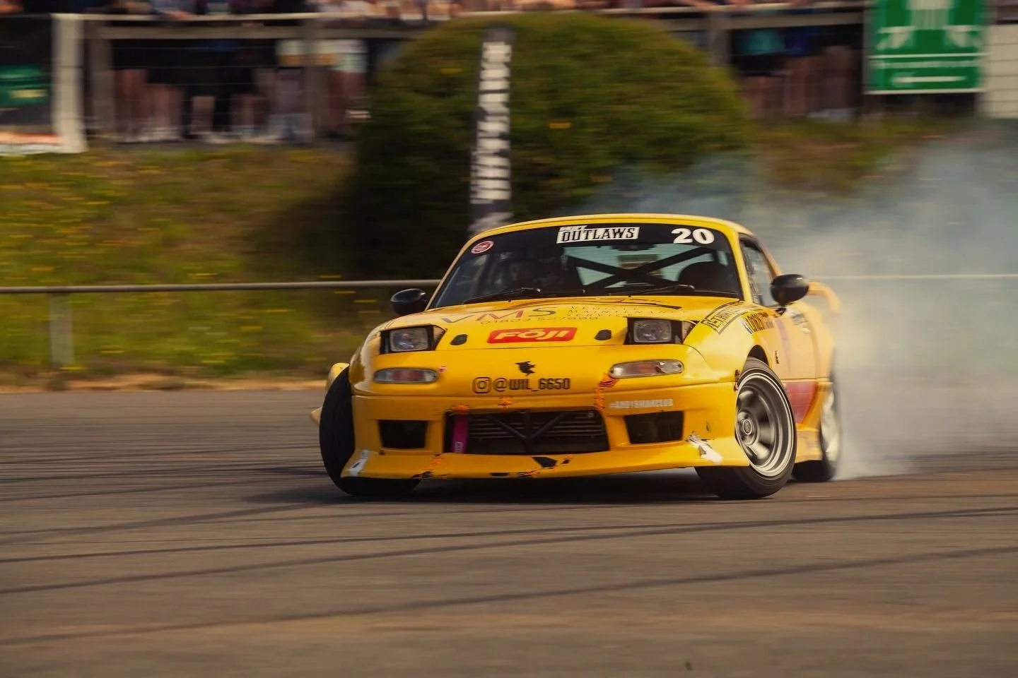 Yellow Mazda Mx5 Miata mk1 drifting on a track, kicking up smoke from the rear tires, with the number 20 and decals, moving swiftly.