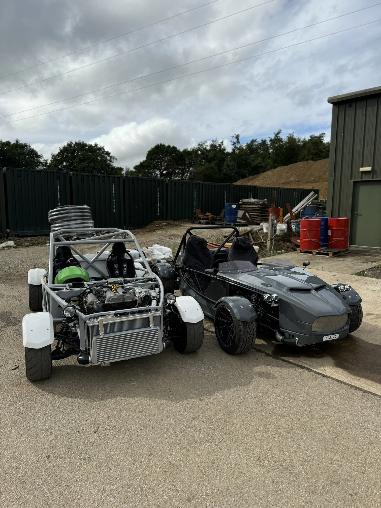 Two high-performance, open-air sports cars parked side by side outdoors on a dirt lot, with construction materials and a green metal building in the background. MEV Exocet